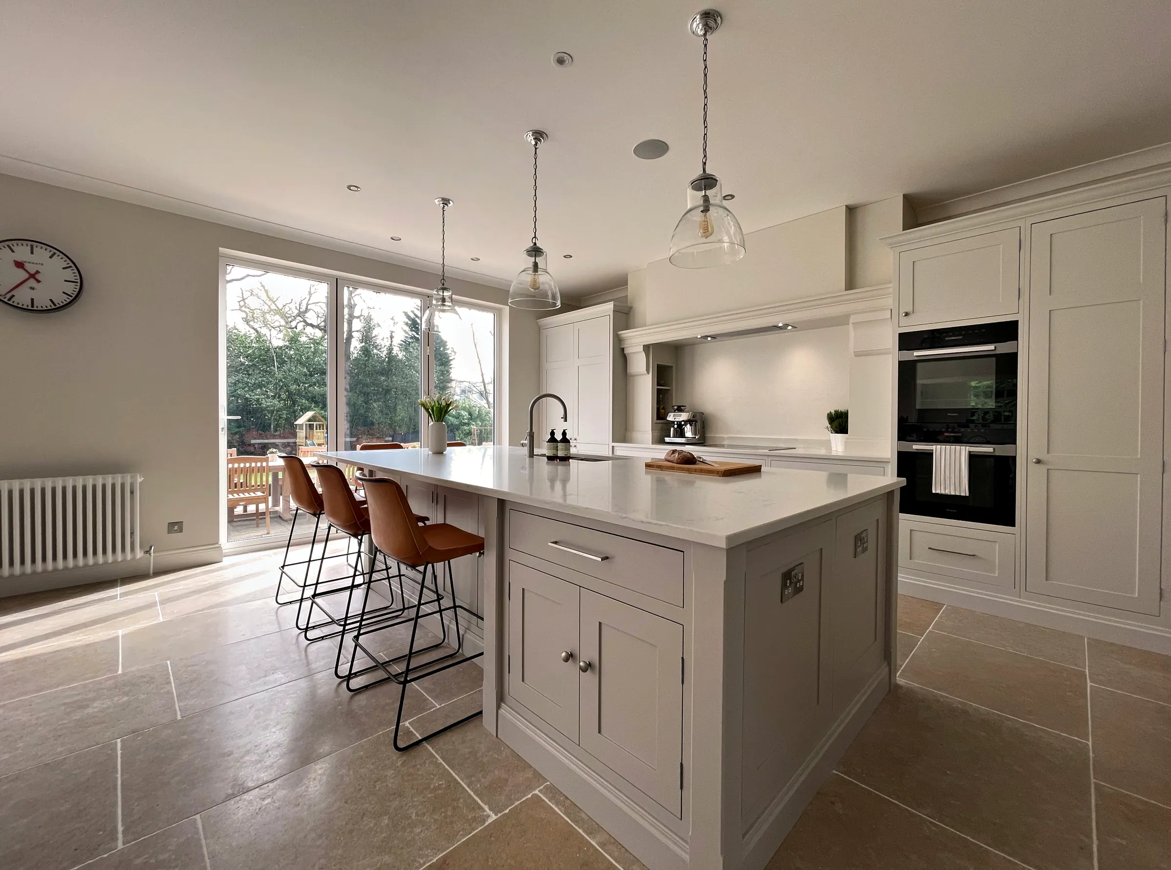 Pale grey painted island with a quartz worktop and leather stools beside the garden doors
