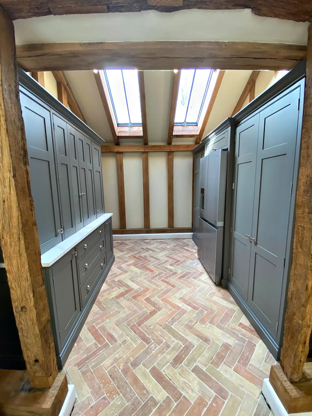 Corridor lined with tall dark taupe cupboards under the oak frame and roof lights on a herringbone brick floor
