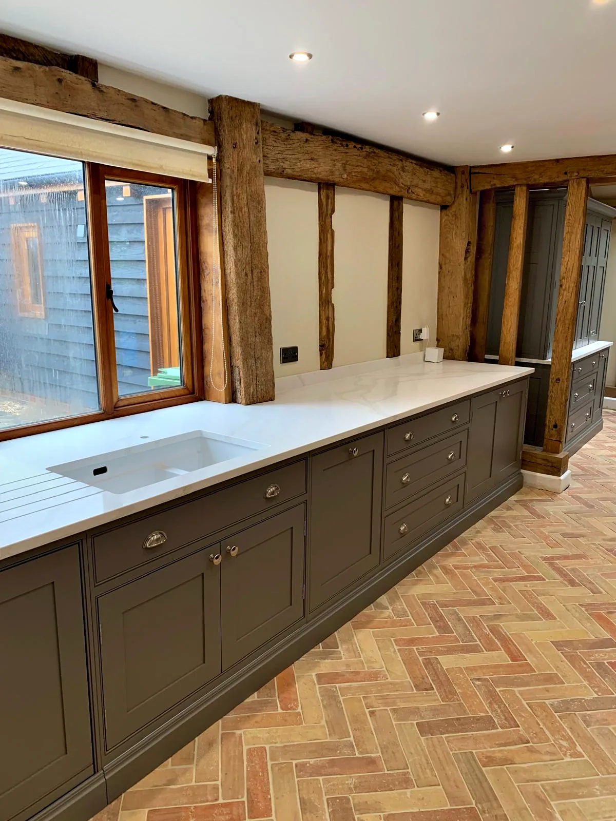 Dark taupe sink run with an undermount sink in a white quartz worktop below a window and the oak frame