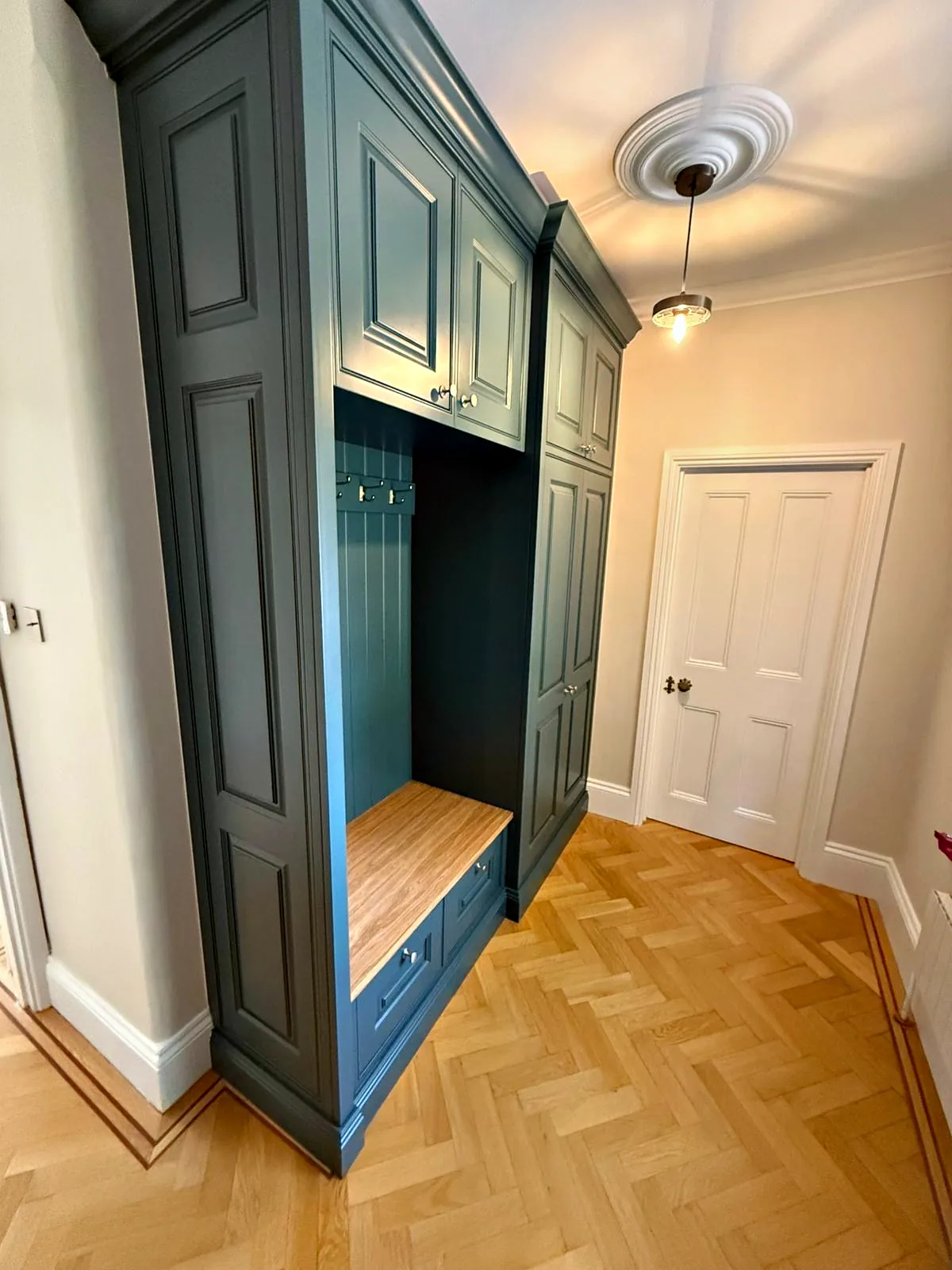 Deep teal hall unit at Chesham House with an oak seat, coat hooks and tall cupboards on herringbone parquet