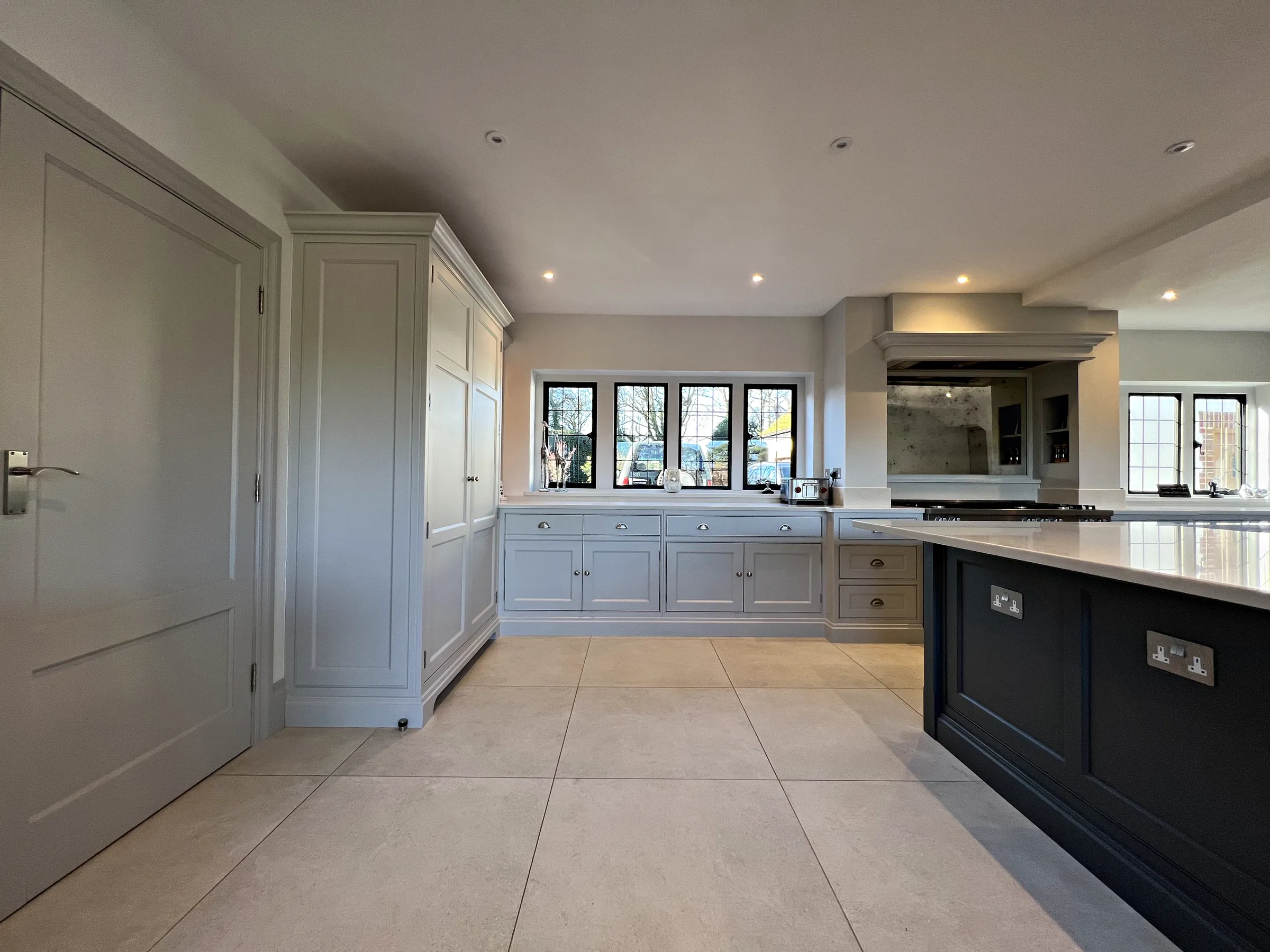 Tall pale grey larder cupboard and the wall run beneath leaded windows, with the end of the Downpipe island in front