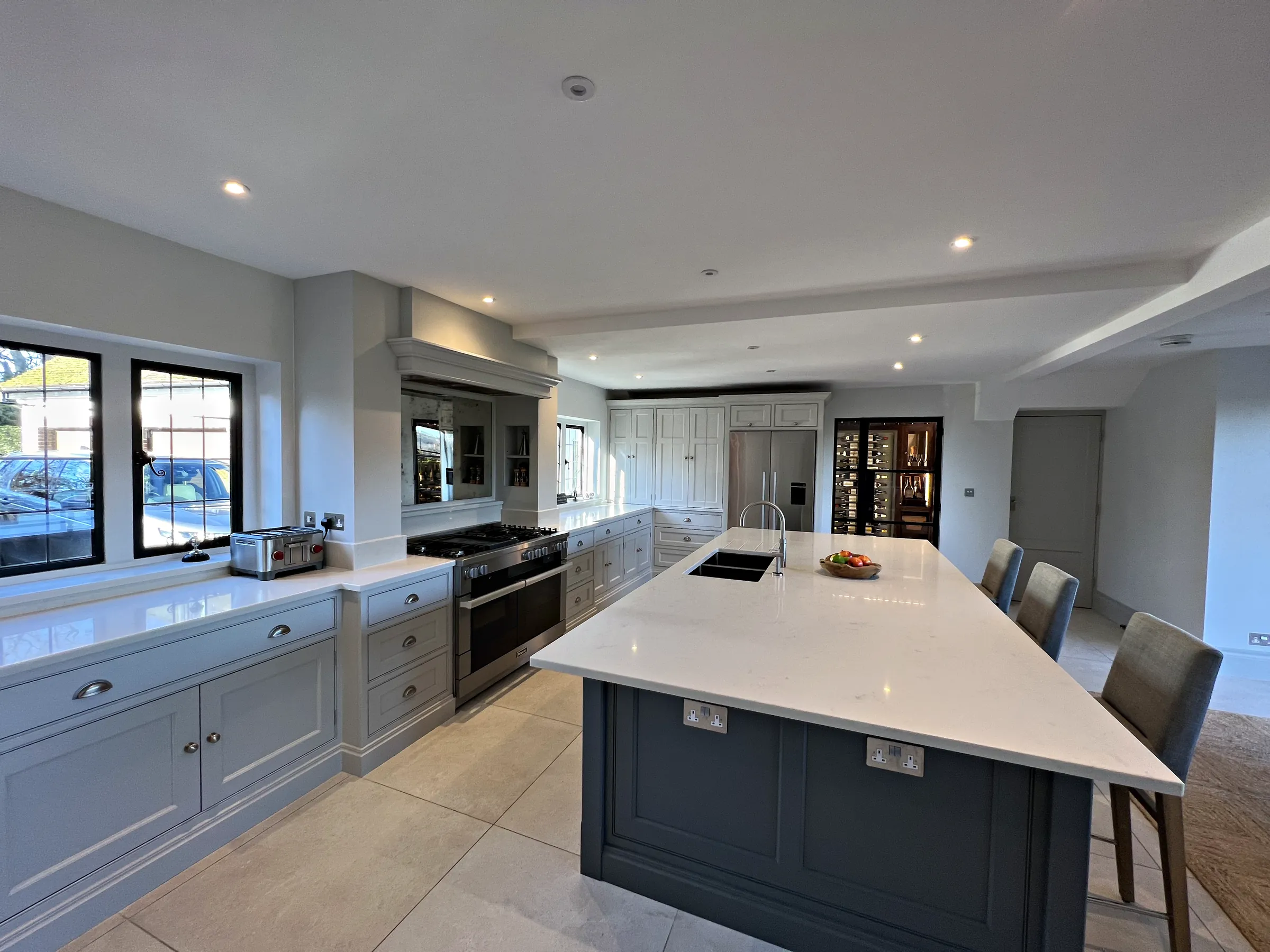 Slate blue island with a sink and bar stools, a pale grey range run and tall units beyond