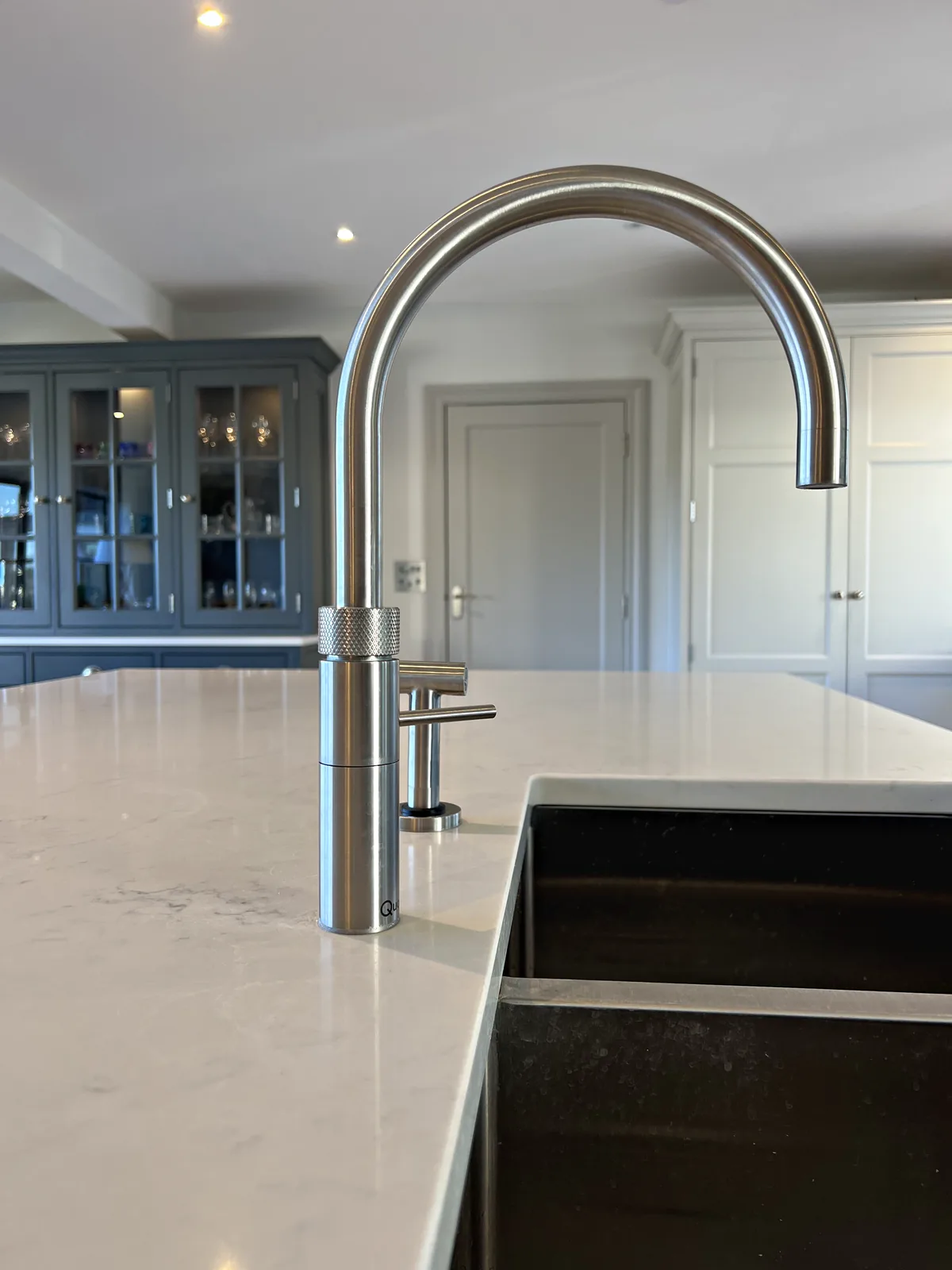 Brushed steel boiling water tap over an island sink with a glazed dresser beyond