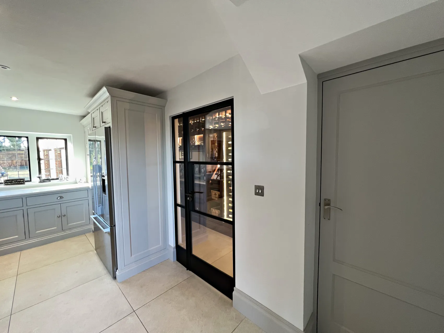 Hallway with a black steel glazed wine room door beside a tall pale grey cupboard, the kitchen beyond
