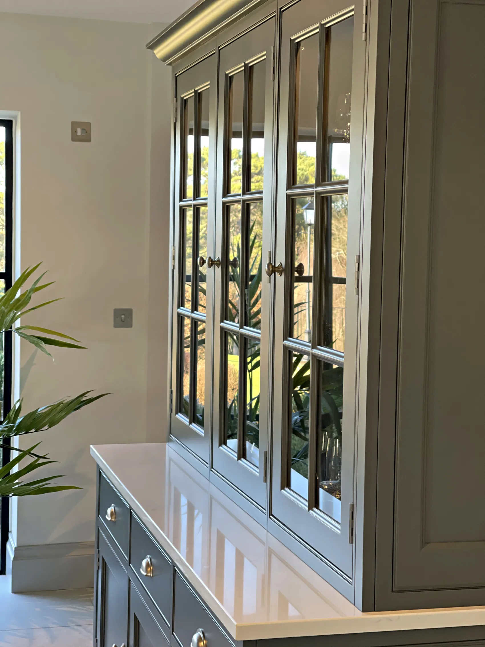 Slate grey glazed dresser with in frame doors beside a palm in a bright room