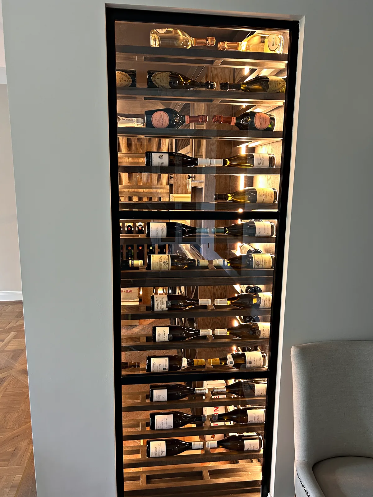 Tall glazed wine cabinet set into a pale wall, lit from within, with bottles resting on dark wood racks