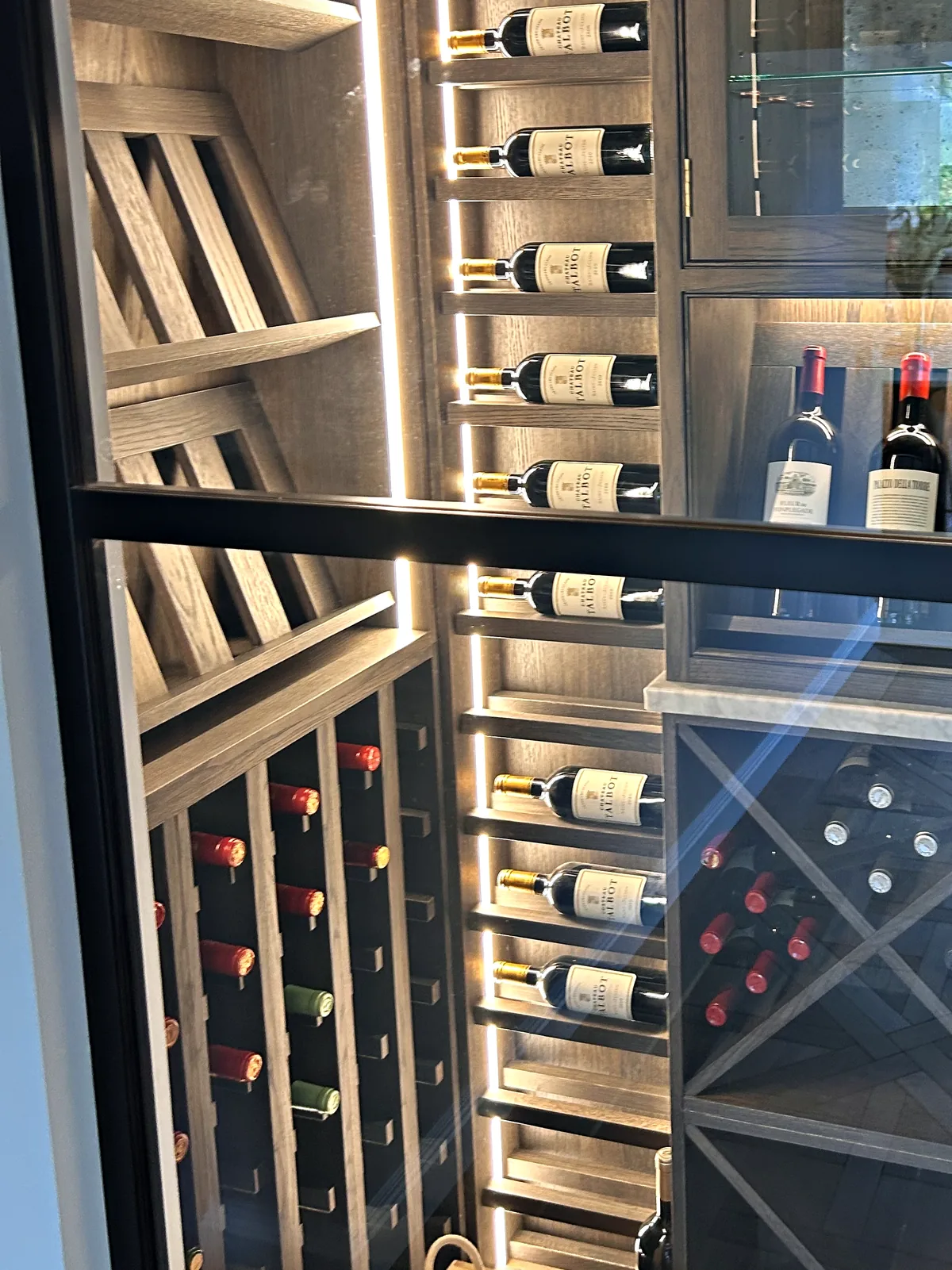 Wine room lined in dark wood behind black framed glazing, with lit bottle racks, a display niche and diamond wine bins