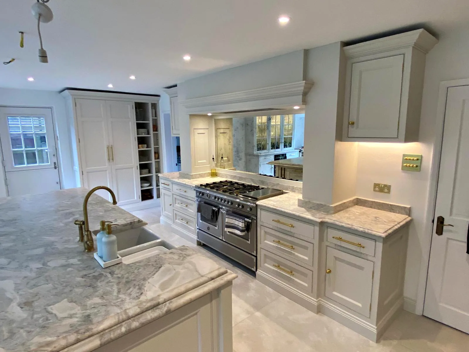 Range wall with a mirrored splashback under a painted canopy, seen past a brass tap at the island sink