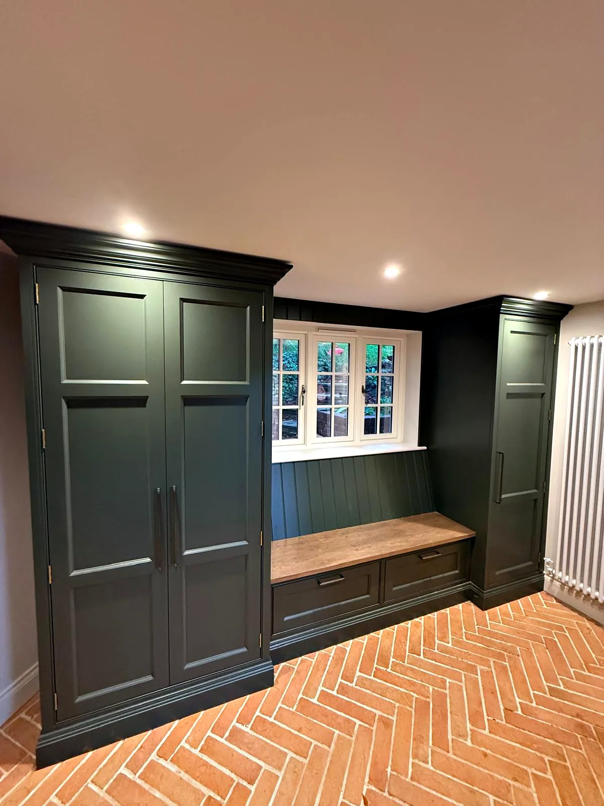 Dark green boot room with tall cupboards, an oak bench and tongue and groove panelling under a window