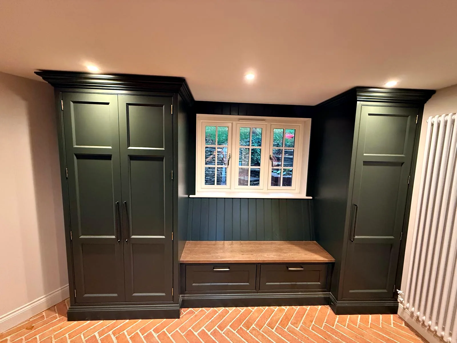 Dark green boot room with two tall cupboards framing an oak bench and tongue and groove panelling under the window
