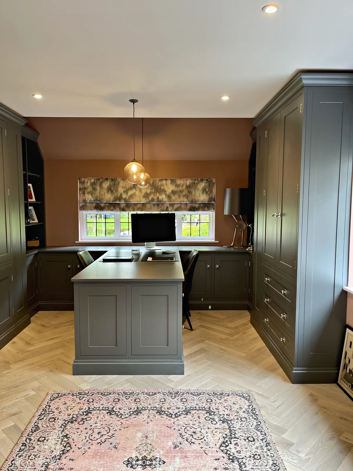 Grey green study with a central desk peninsula, tall cupboards, open bookshelves and a desk run beneath the window