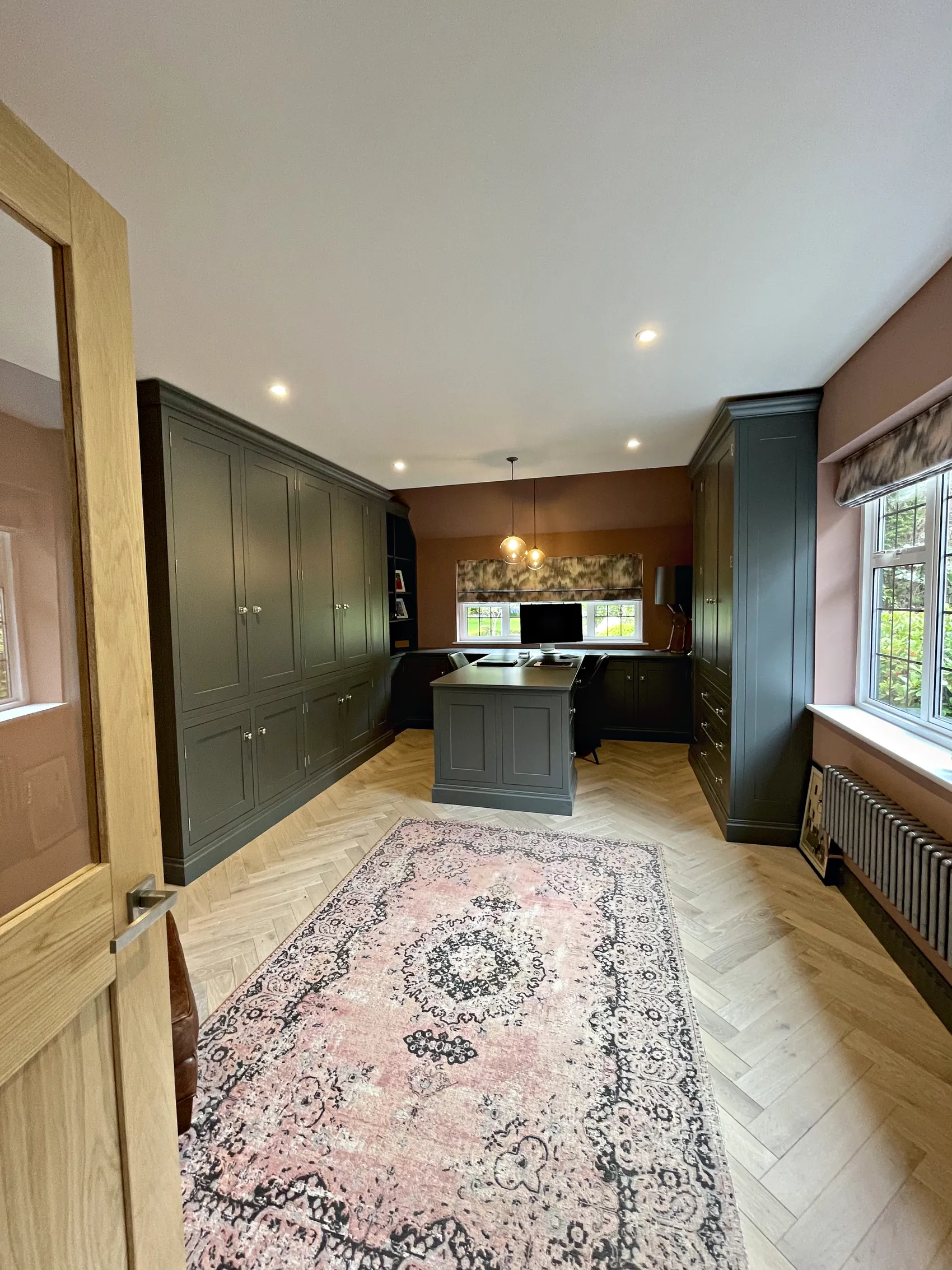Study with a wall of tall grey green cupboards, a desk run under the window and a pink rug