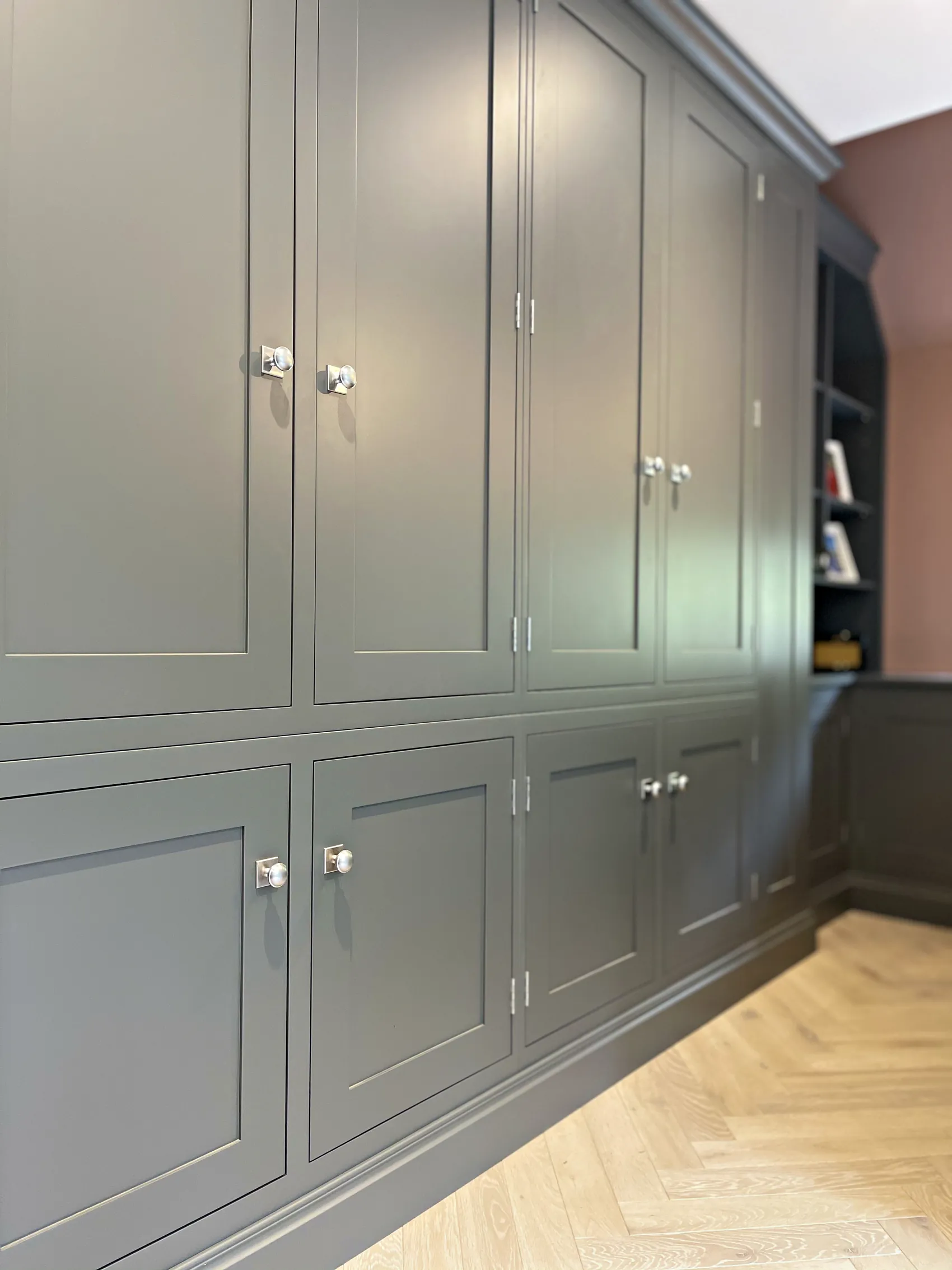 Wall of tall grey green cupboards with nickel knobs running to open bookshelves in a study