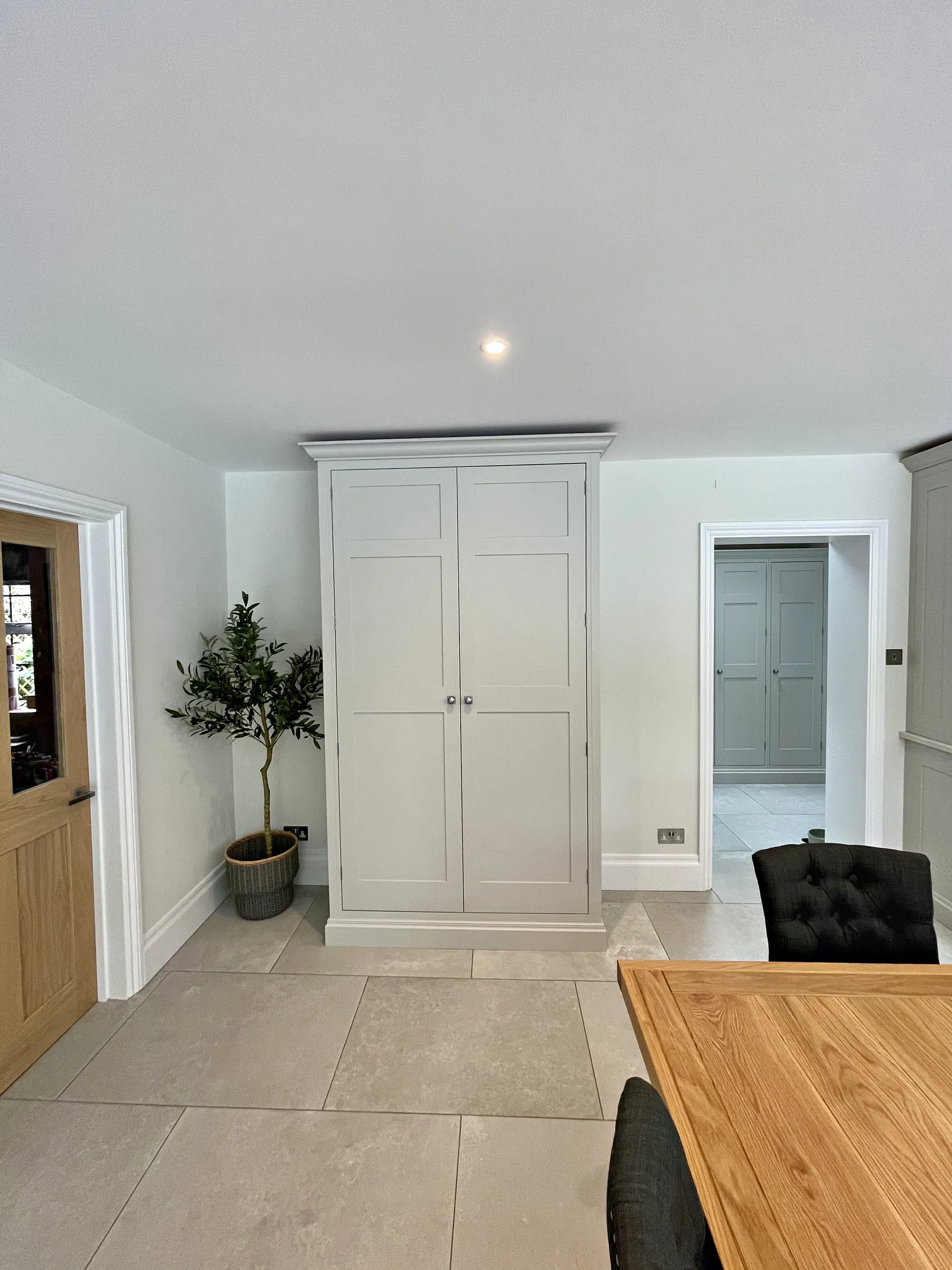 Freestanding pale grey larder cupboard with a cornice beside an olive tree and an oak dining table