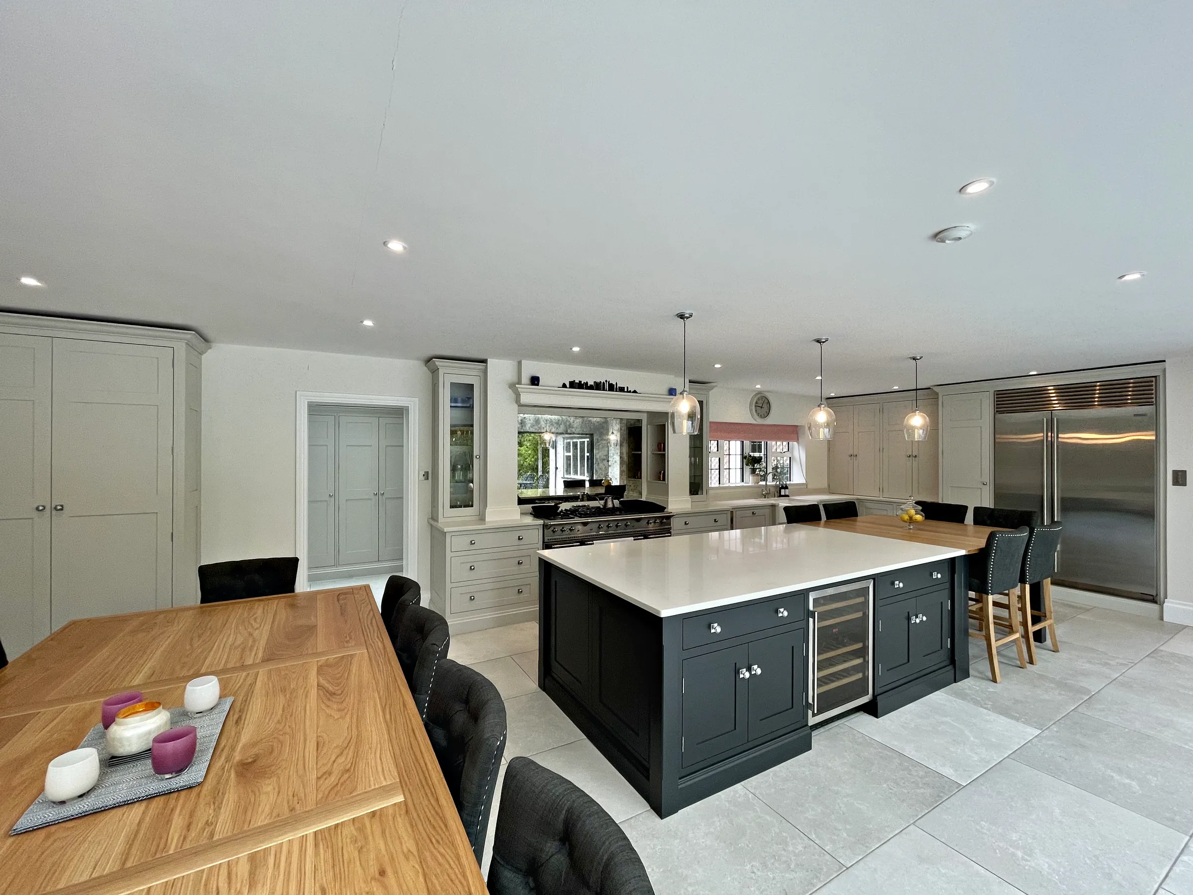 Charcoal island, range canopy, glazed dresser and steel fridge freezer seen past an oak dining table