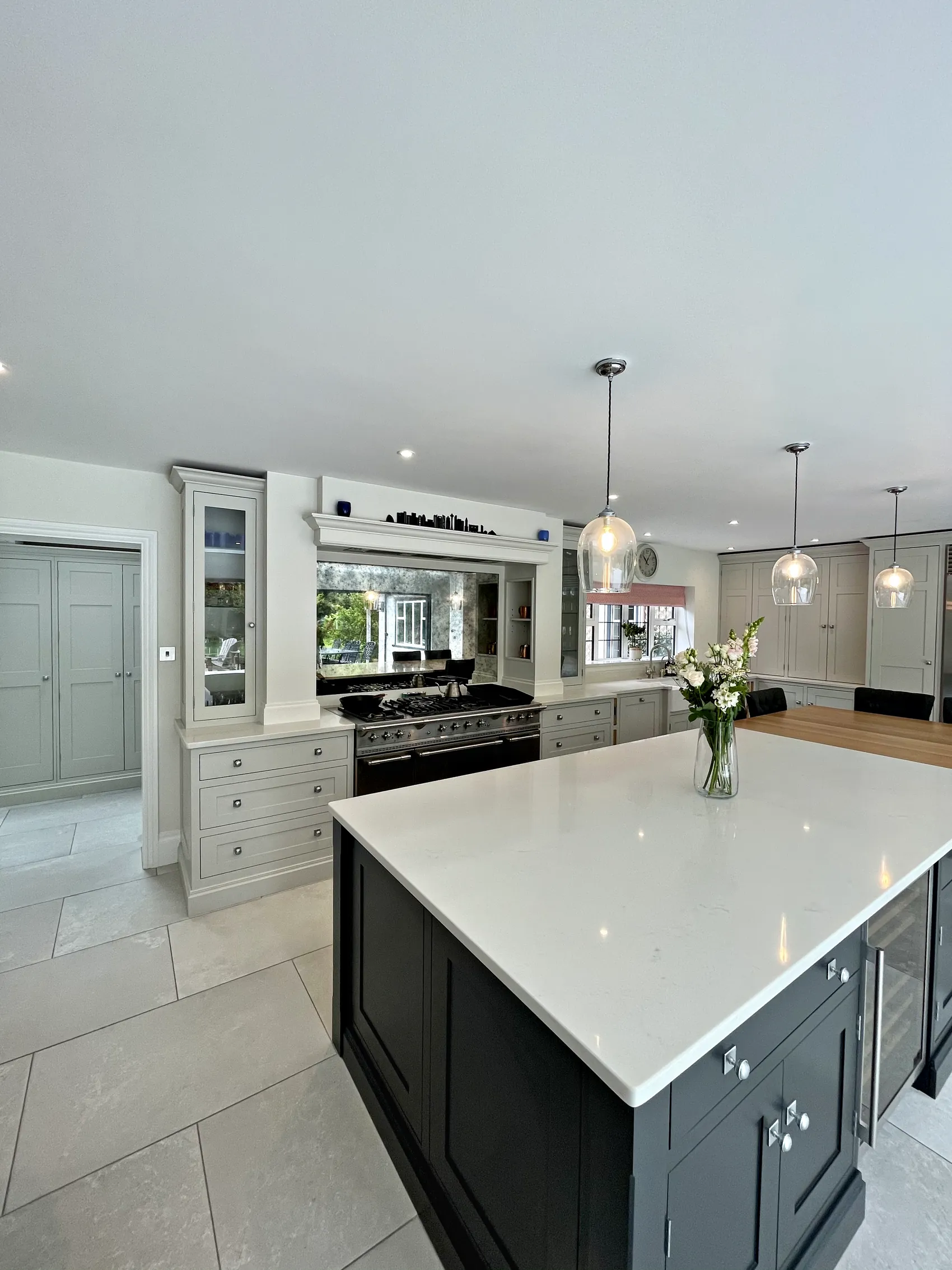 Charcoal island with a white quartz top under glass pendants before a range wall with a glazed dresser