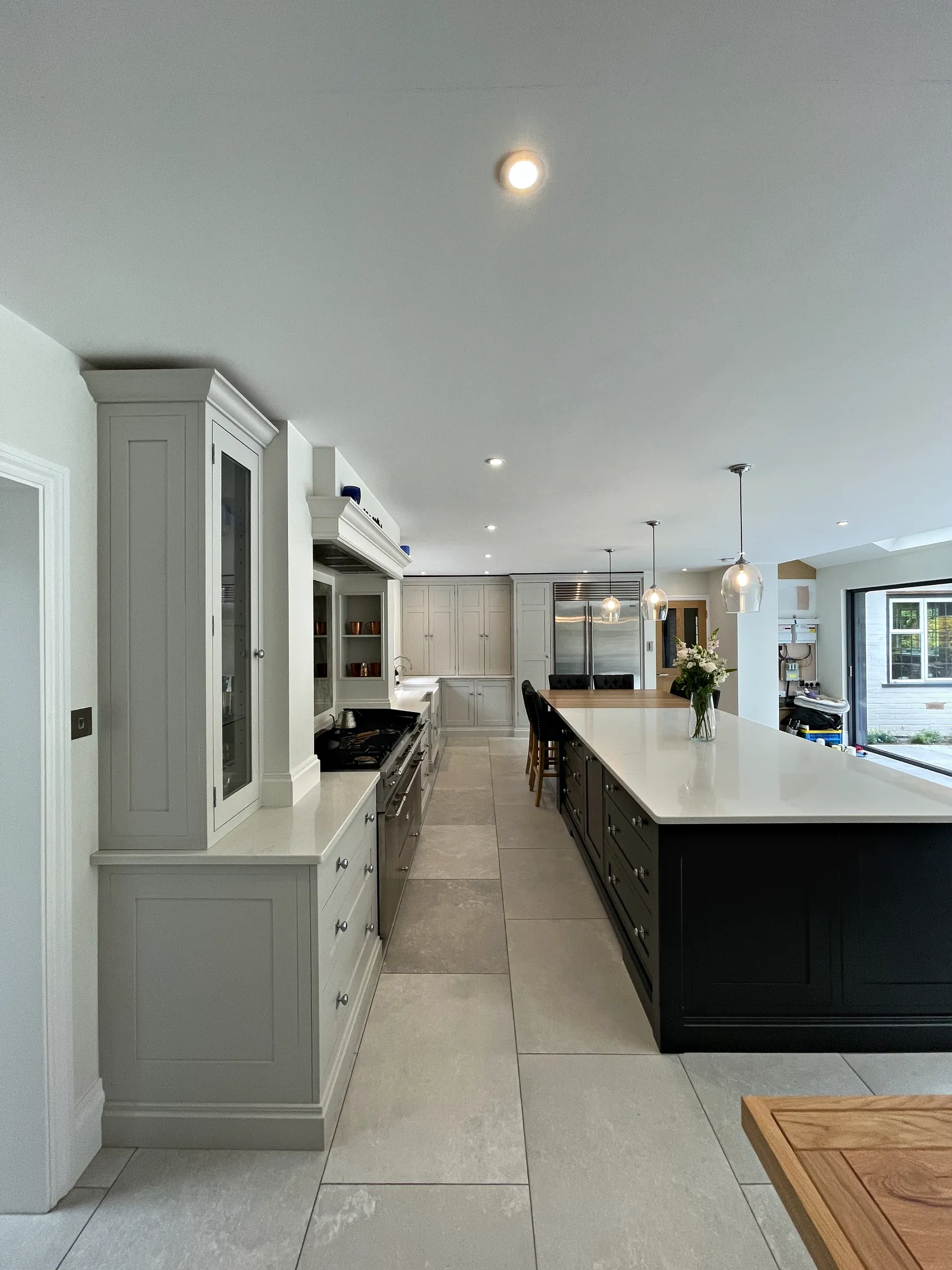 View down a long pale grey kitchen past a glazed dresser to a charcoal island and garden doors