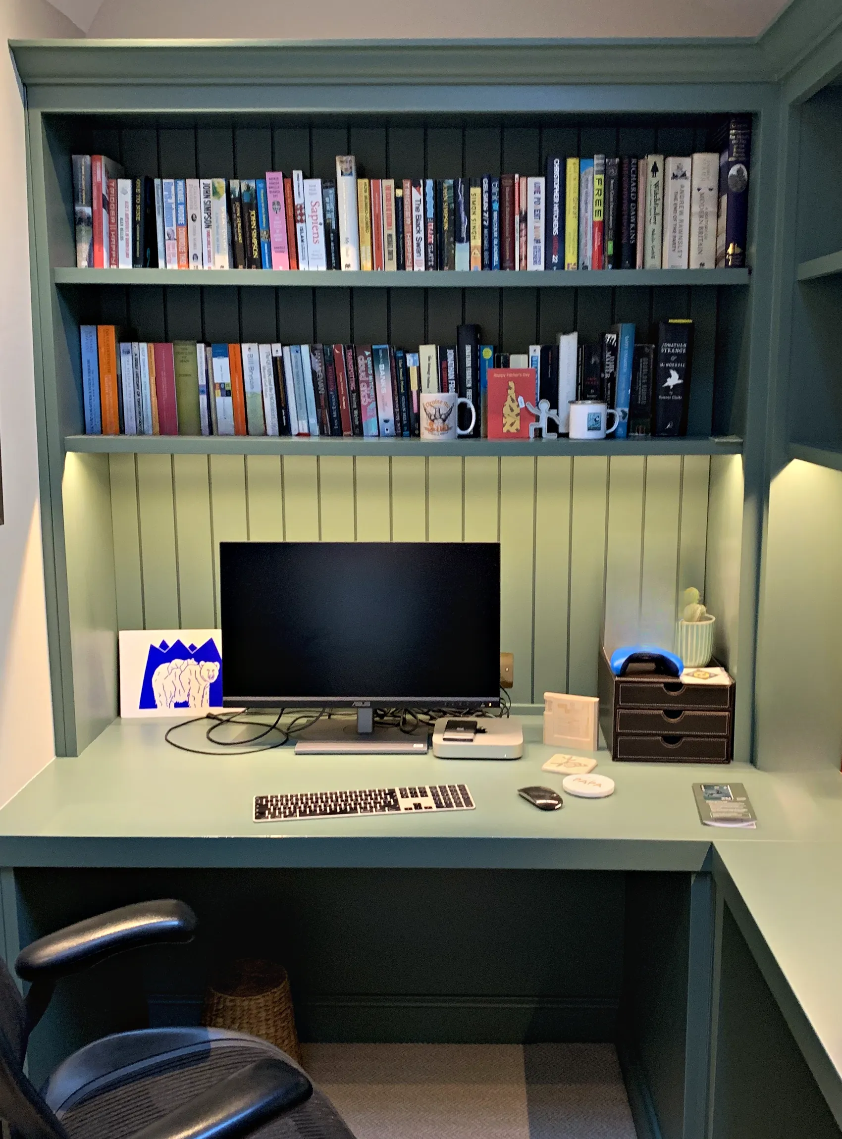 Green painted home office with a built in desk beneath lit open shelving and a tongue and groove panelled back