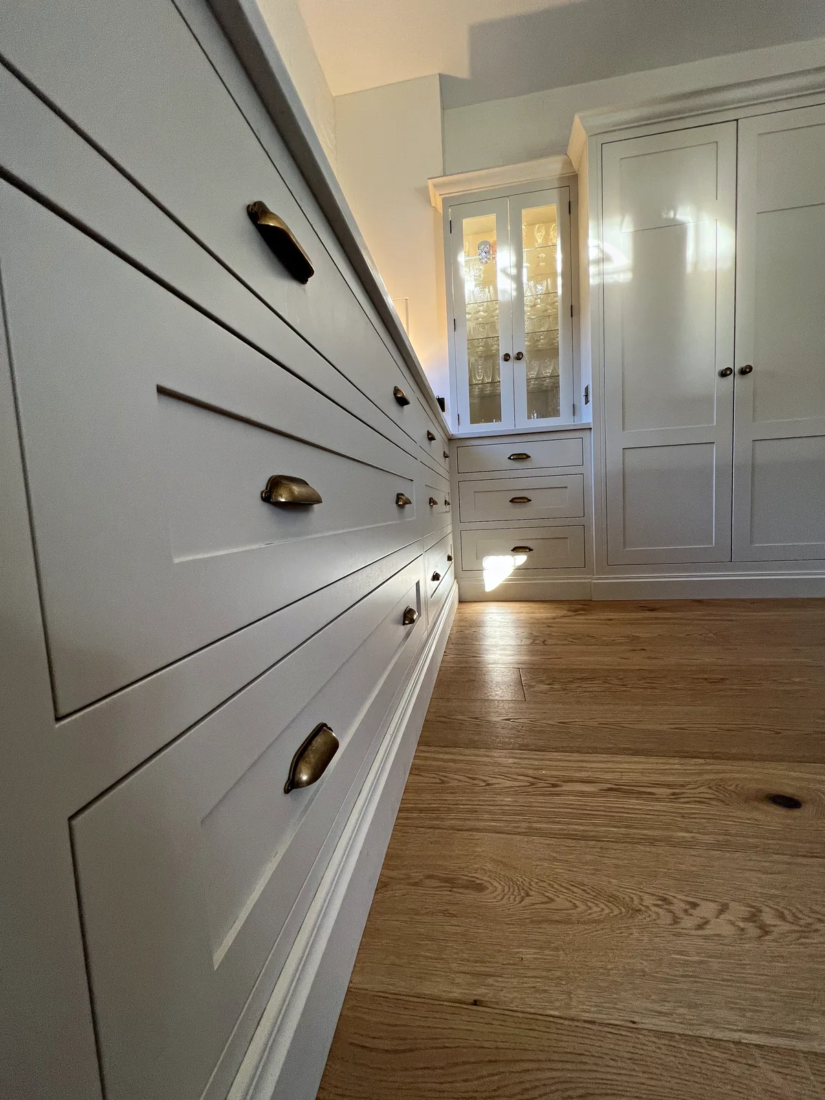 Run of cream drawers with brass cup handles leading to a glazed cabinet and tall cupboard