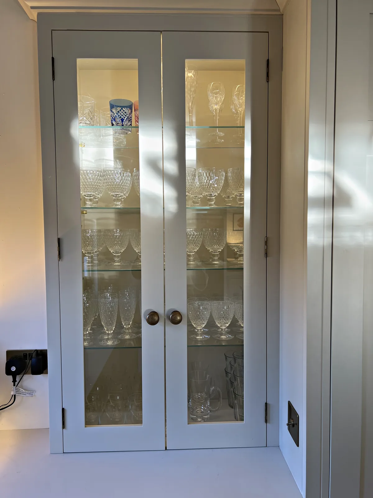 Pale painted glazed cabinet doors with bronze knobs and cut glassware on glass shelves inside