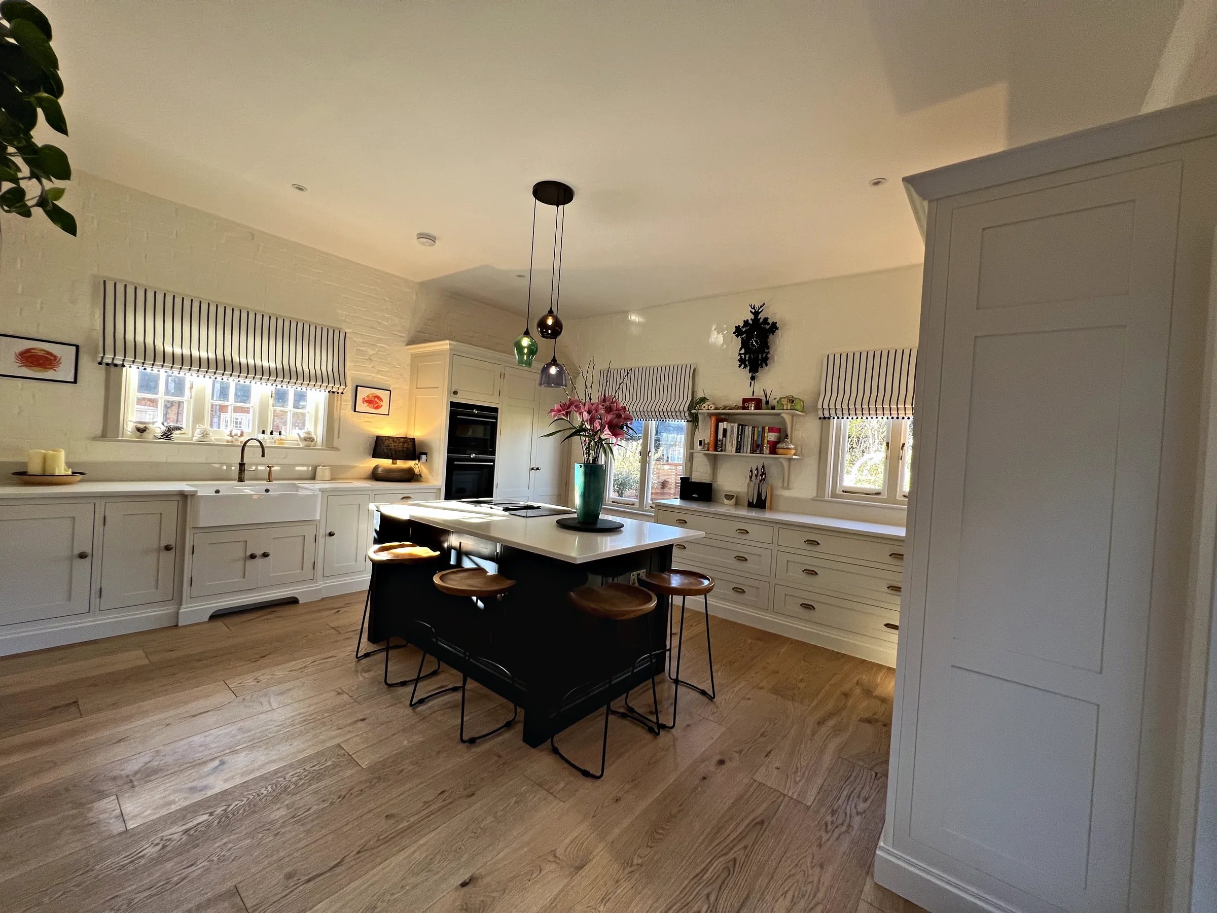 Cream kitchen with a navy island and stools, a Belfast sink run under striped blinds and a tall cupboard