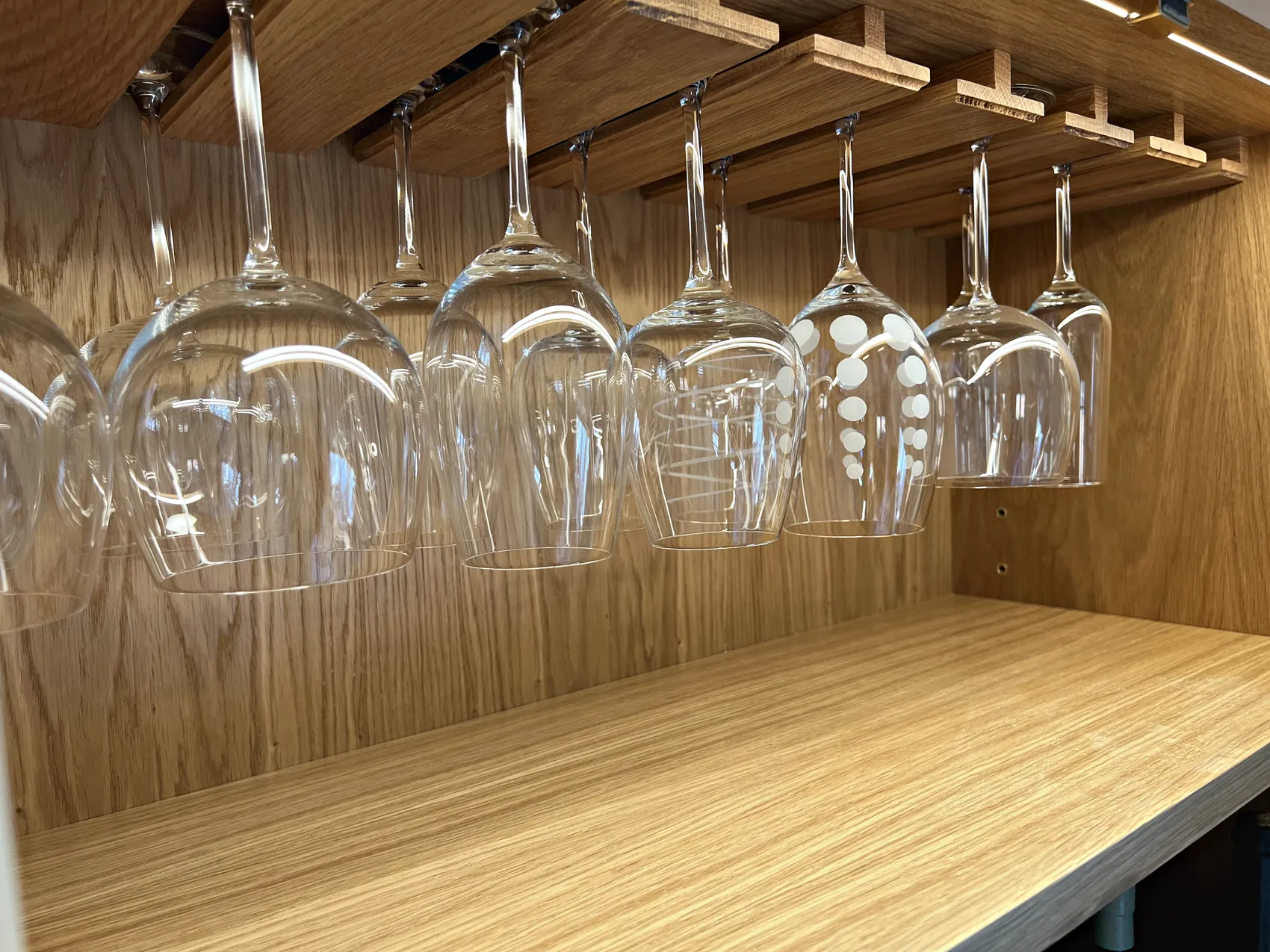 Oak glass rack holding upturned wine glasses above an oak shelf inside a drinks cupboard