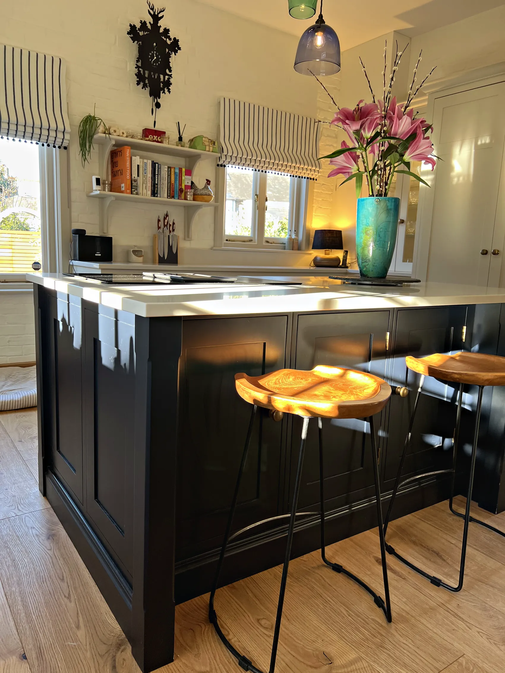 End of a navy island with wooden saddle stools, cream cabinetry and bracket shelves beyond