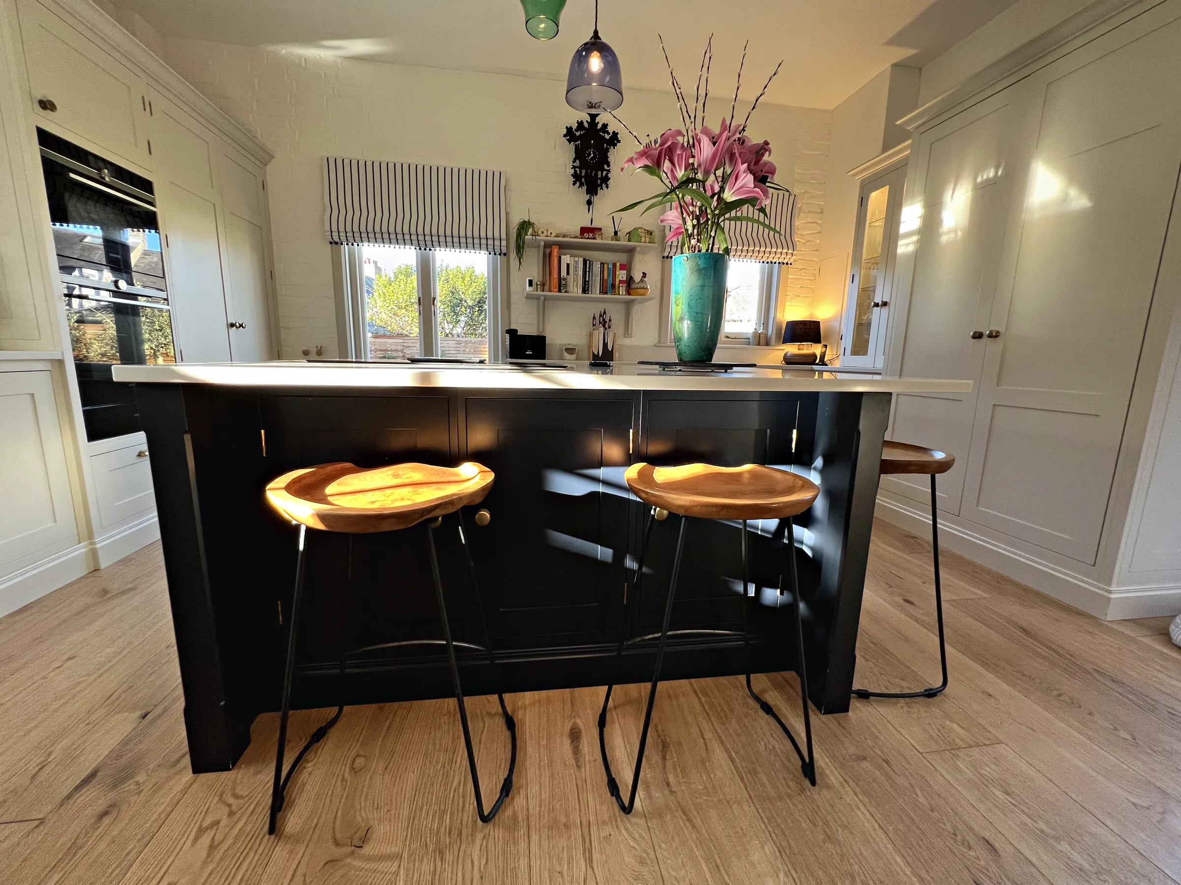 Navy island with wooden saddle stools on a wide oak floor, built in ovens and tall cream cupboards