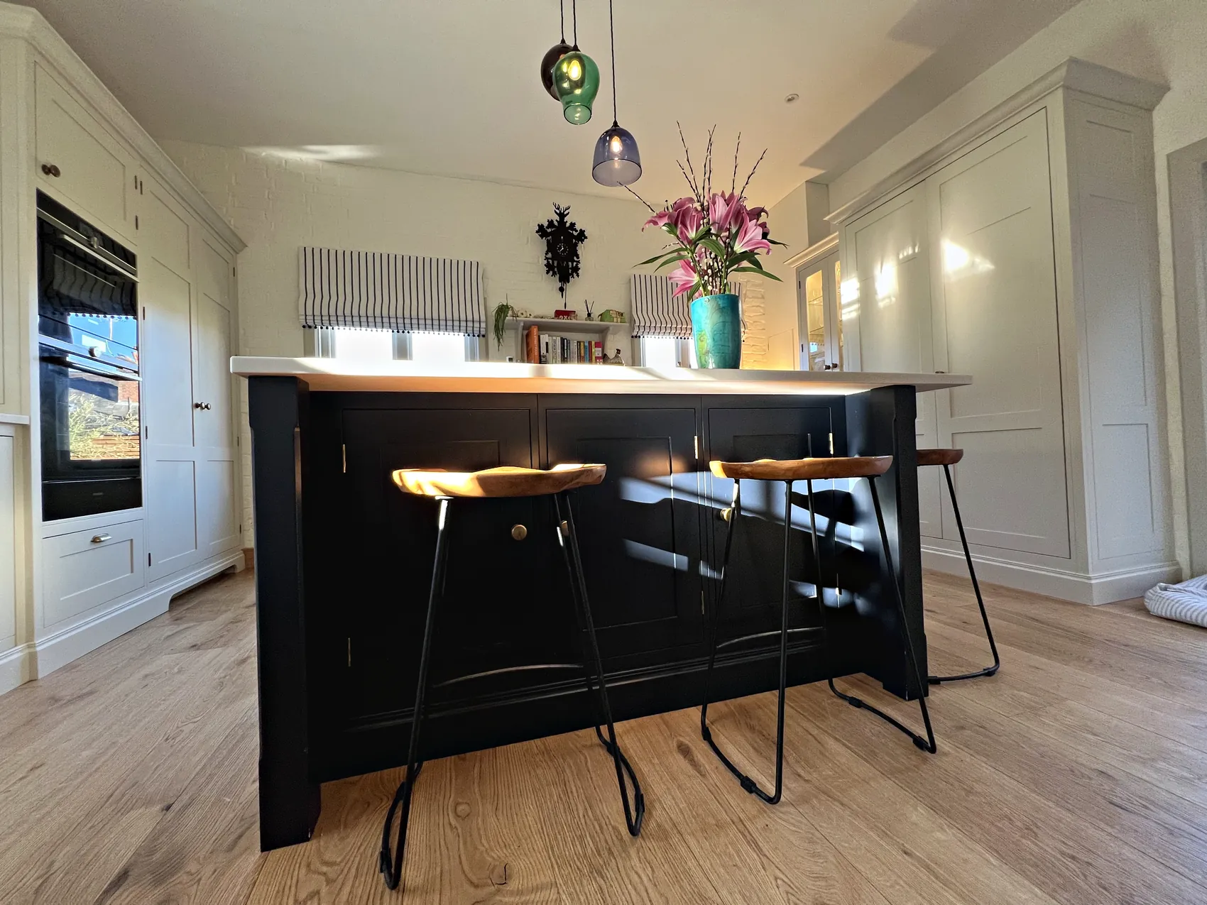 Navy island with wooden saddle stools in front of a cream sink run under striped blinds on an oak floor