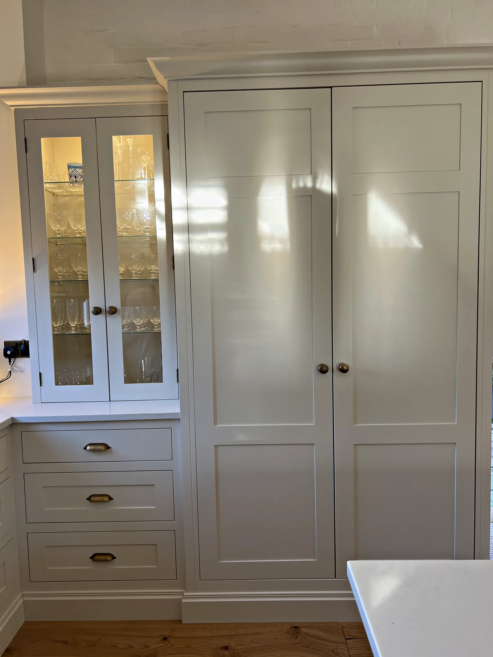 Glazed cream cabinet over drawers with brass cup handles beside a tall cream double cupboard