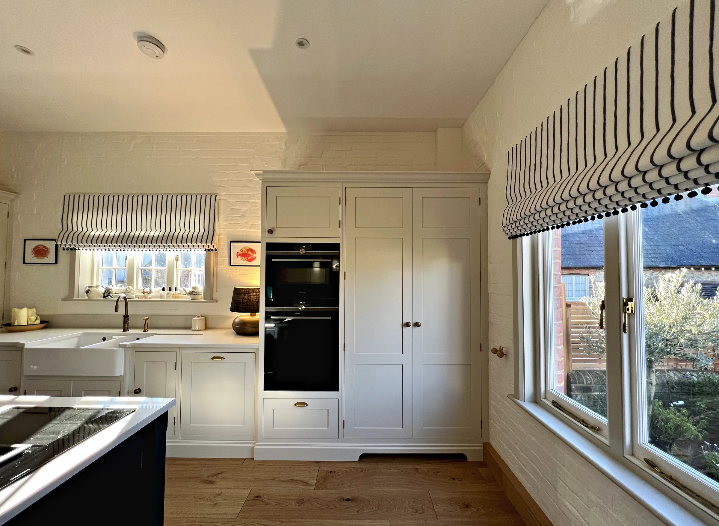 Tall cream cupboards with built in ovens beside a garden window, a Belfast sink run on the painted brick wall