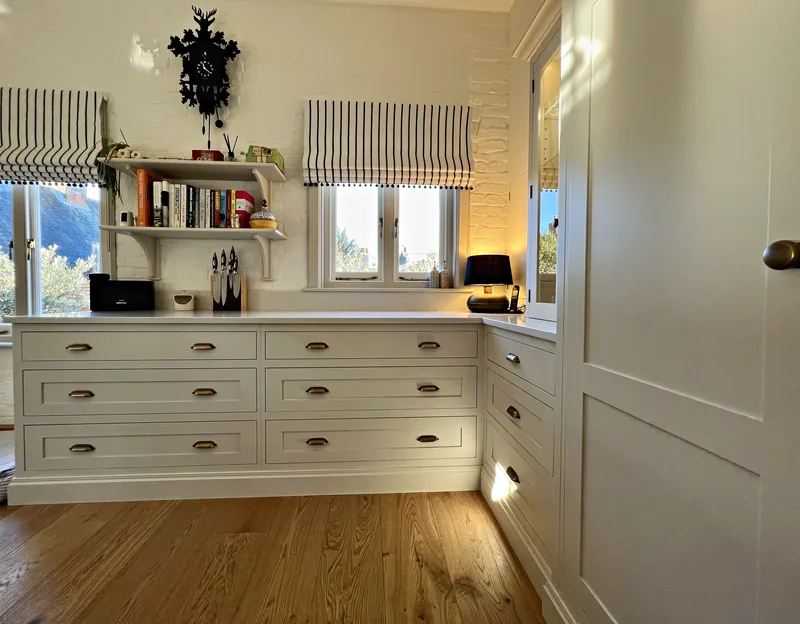 Long run of drawers with antique brass cup handles under bracket shelves between two windows