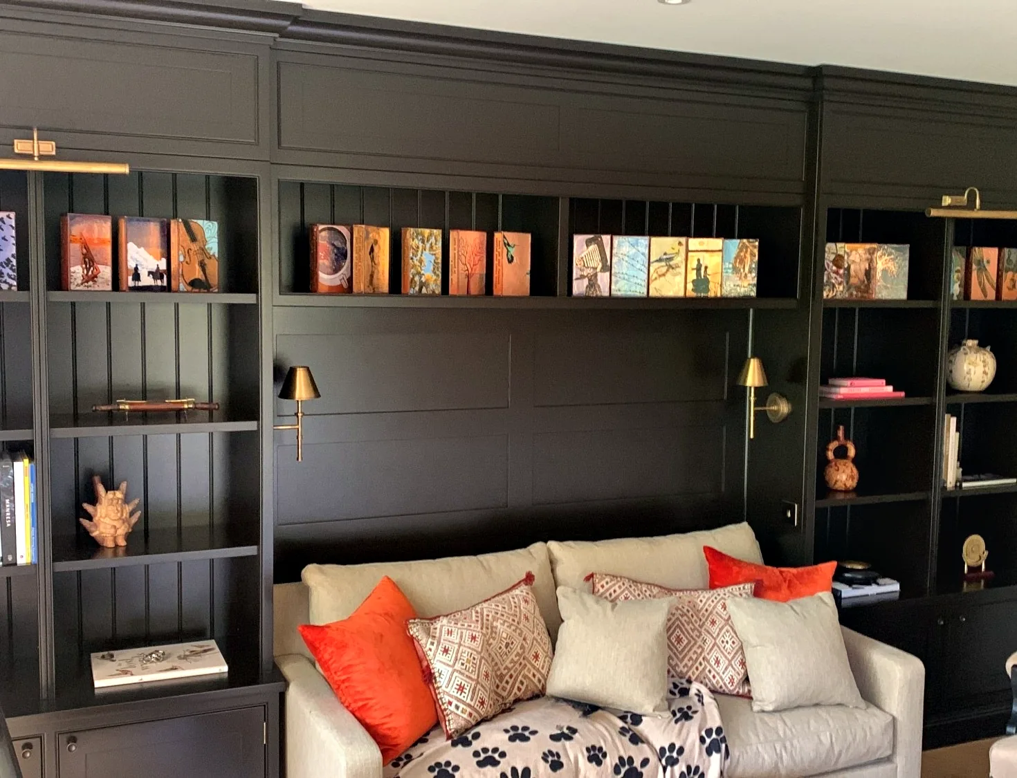 Living room library wall at Roughwoods, painted in a very dark colour, with tongue and groove backed shelves, brass picture lights and wall lights