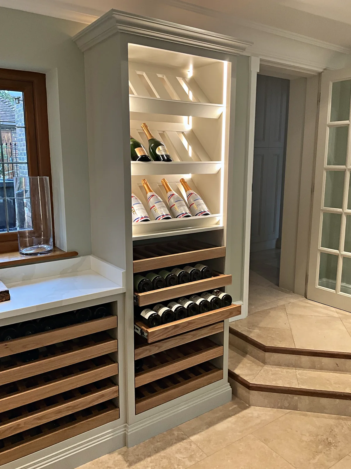 Lit wine display tower with angled shelves above an oak pull out bottle drawer in sage grey cabinetry