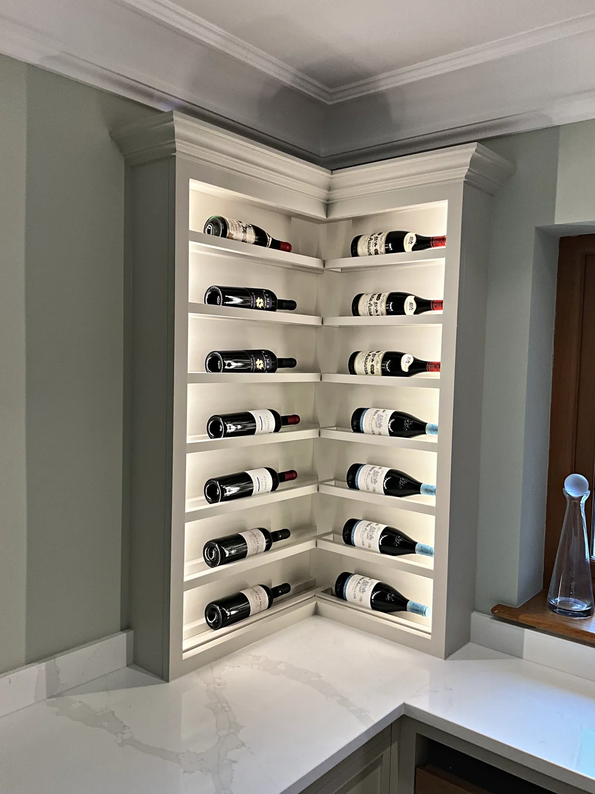 Painted corner wine display cabinet with lit shelves holding bottles on their sides above a white veined worktop