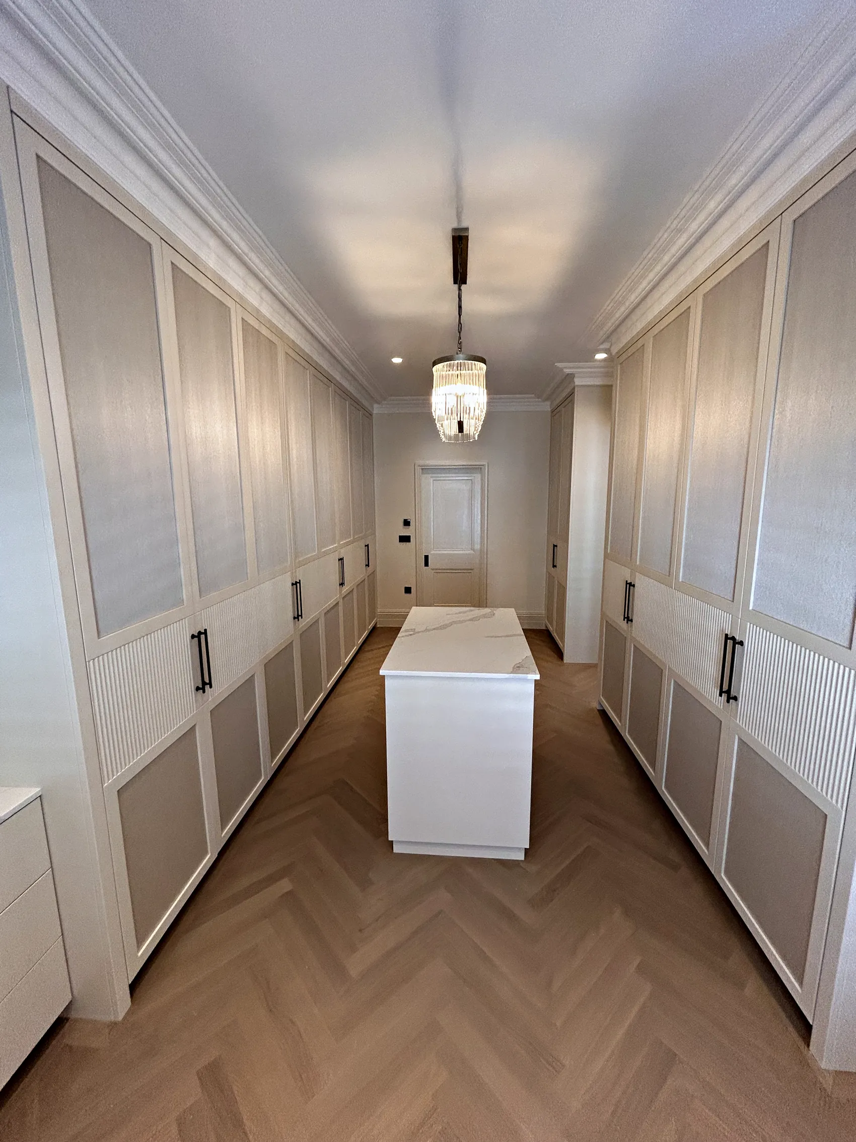 Walk in dressing room with cream wardrobes on both sides, reeded and textured panels and a central island under a chandelier