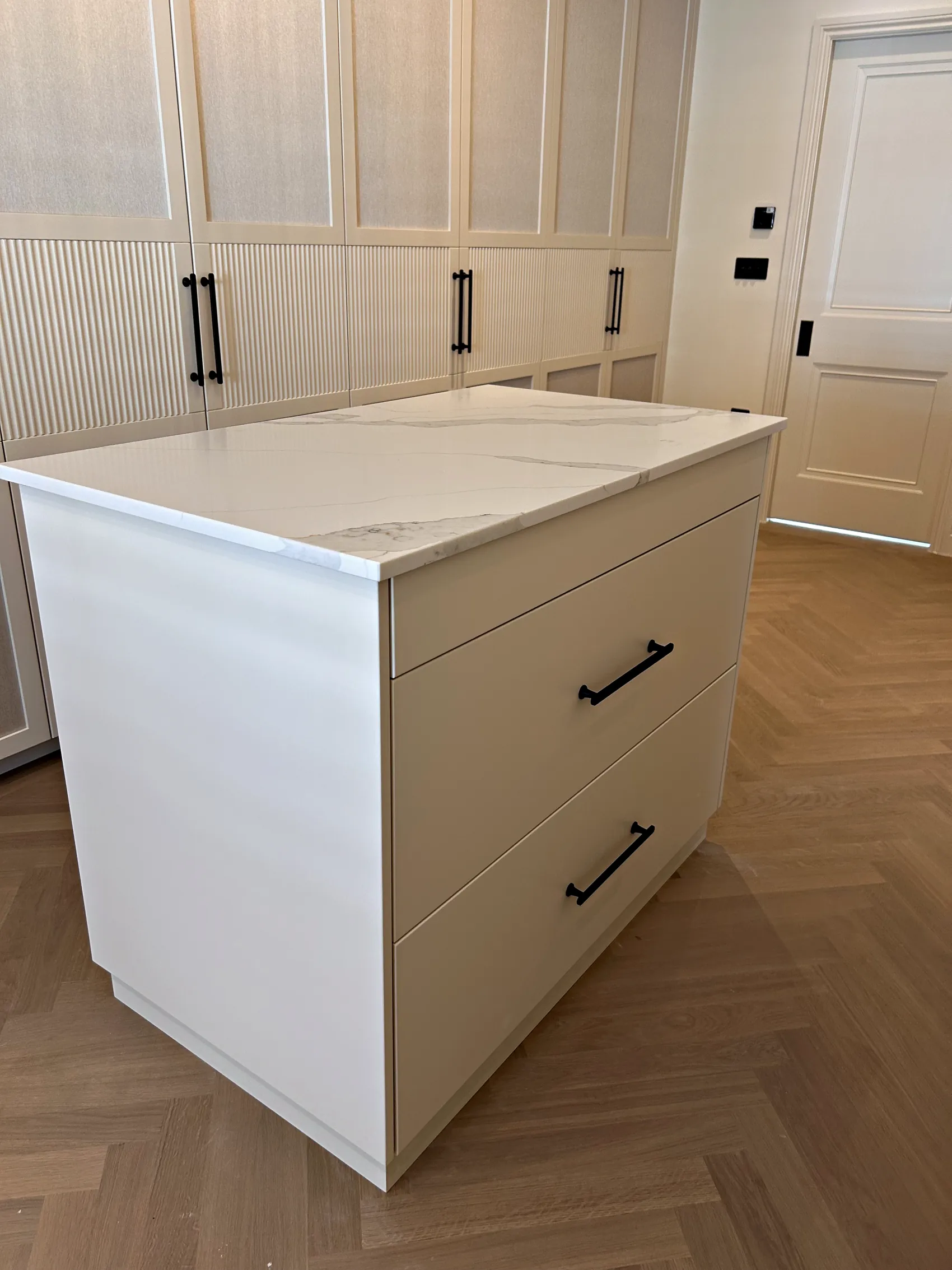 Cream dressing island with three drawers, black bar handles and a white veined top in a walk in dressing room