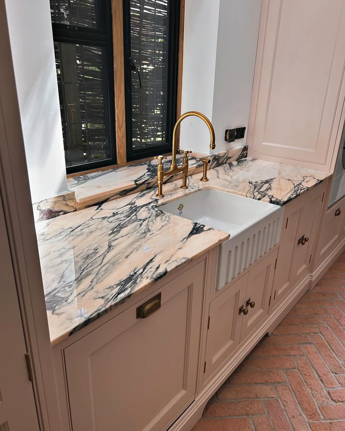 Ribbed Belfast sink in a veined marble worktop with a brass bridge tap over blush pink cabinetry