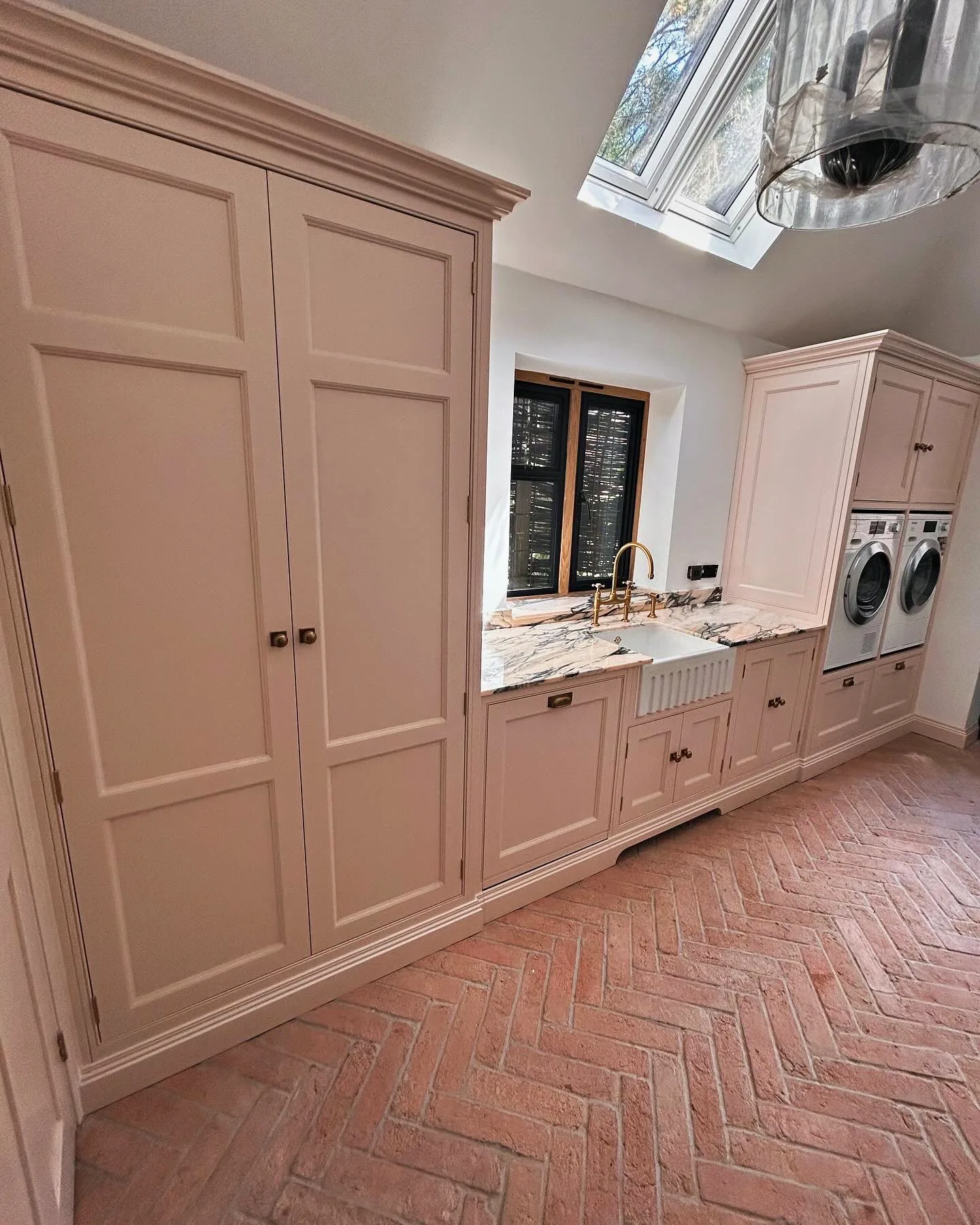 Blush pink utility with a tall double cupboard, a Belfast sink and stacked appliances on a terracotta floor