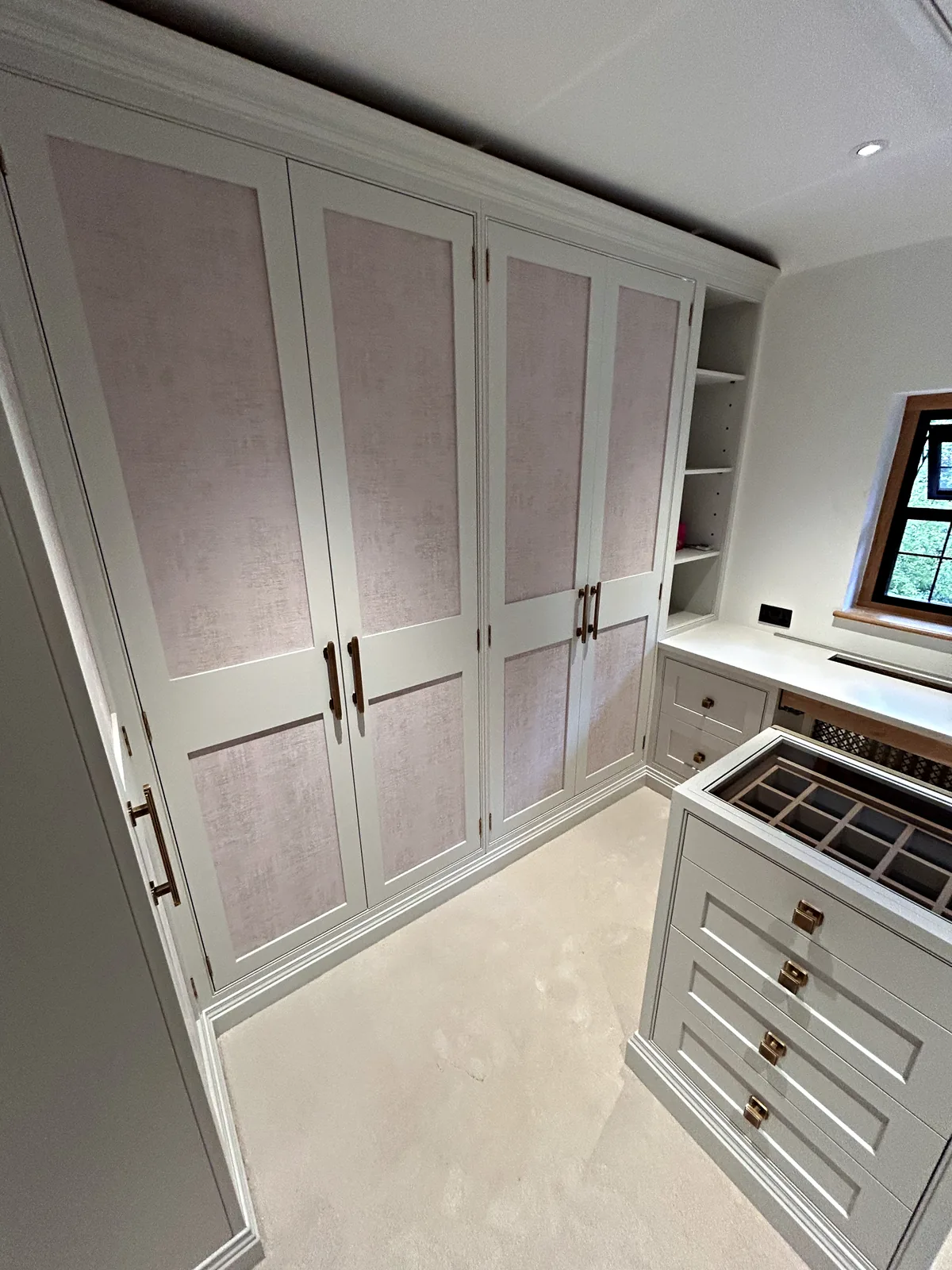 Dressing room wardrobes with blush fabric panels and bronze handles beside a drawer island with a glass topped tray