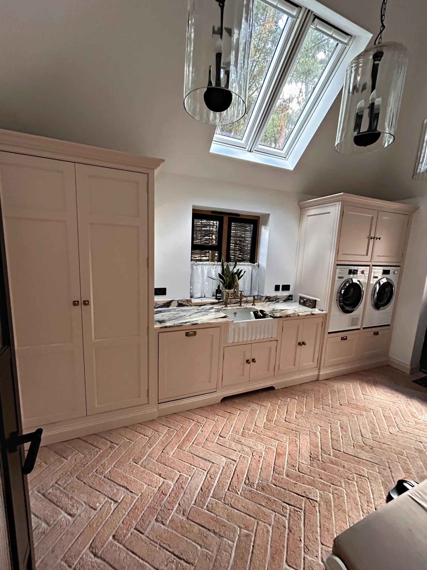 Blush pink utility under a roof window with a tall cupboard, a Belfast sink and a stacked washer and dryer on a terracotta herringbone floor