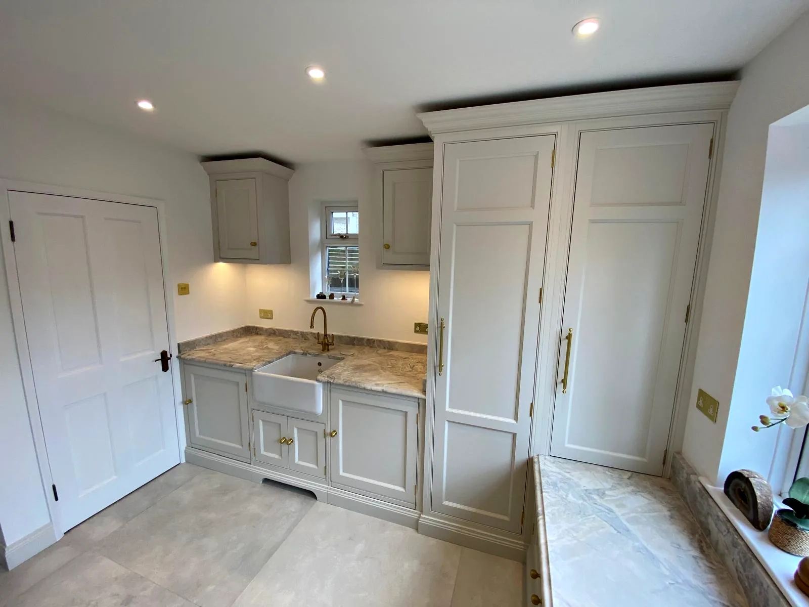 Painted back kitchen with a Belfast sink, a brass tap, veined stone worktops, tall cupboards and a window seat