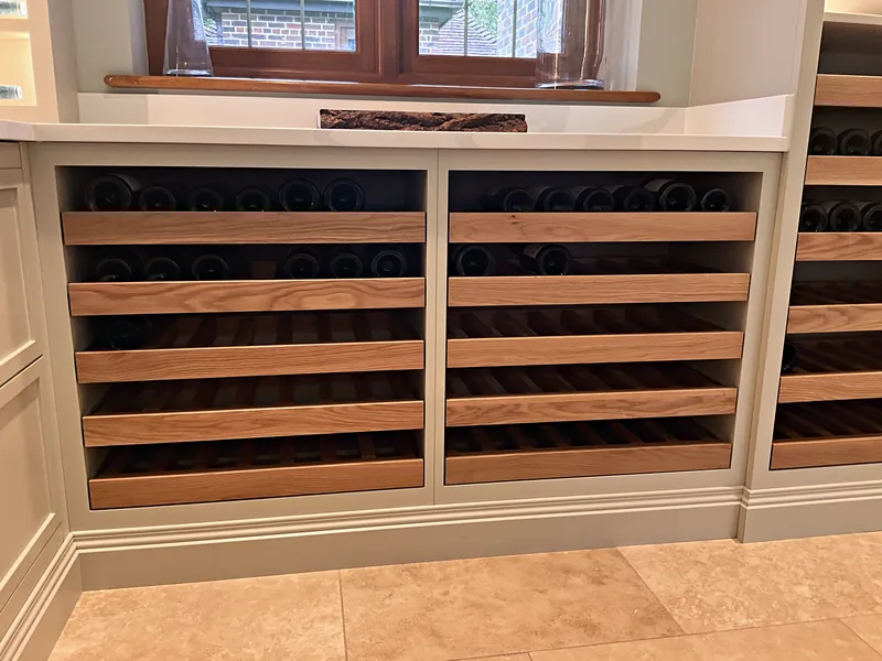 Oak slatted pull out wine trays holding bottles in pale sage grey base cupboards beneath a window