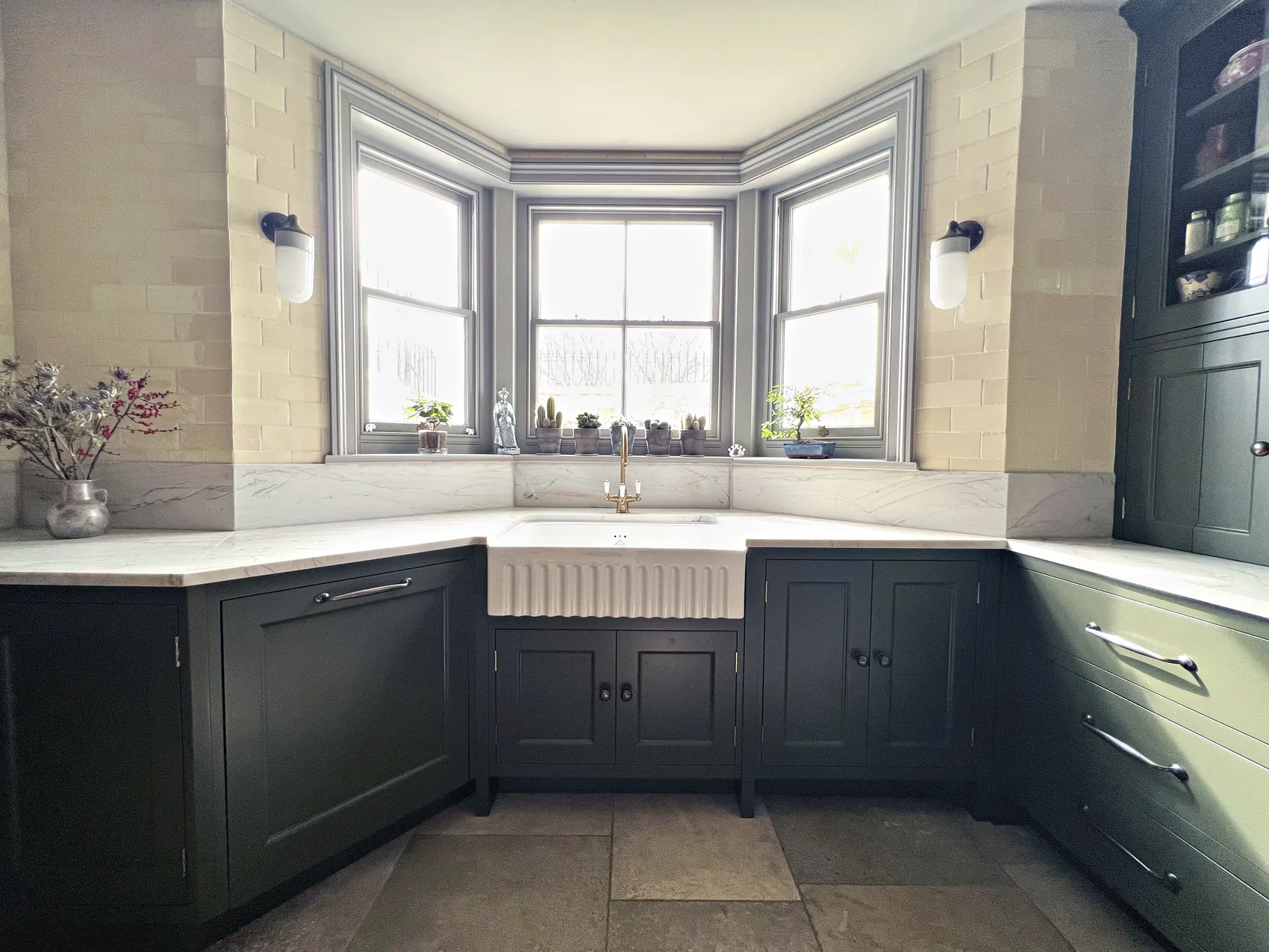 Olive green kitchen around a bay window with a fluted Belfast sink, a brass tap and white veined worktops