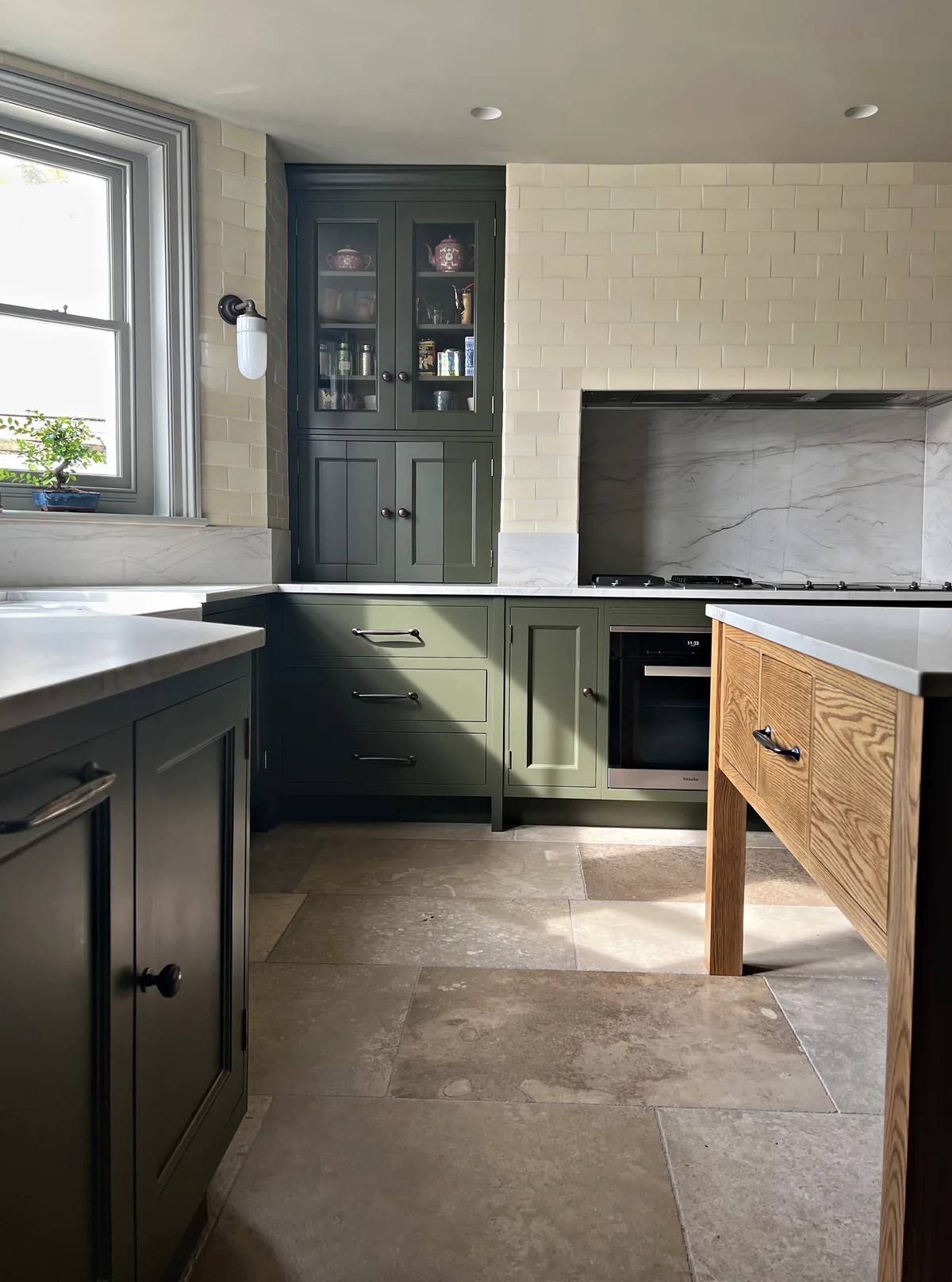 Olive green kitchen with a glazed wall cabinet, a stone splashback behind the hob and an oak table island