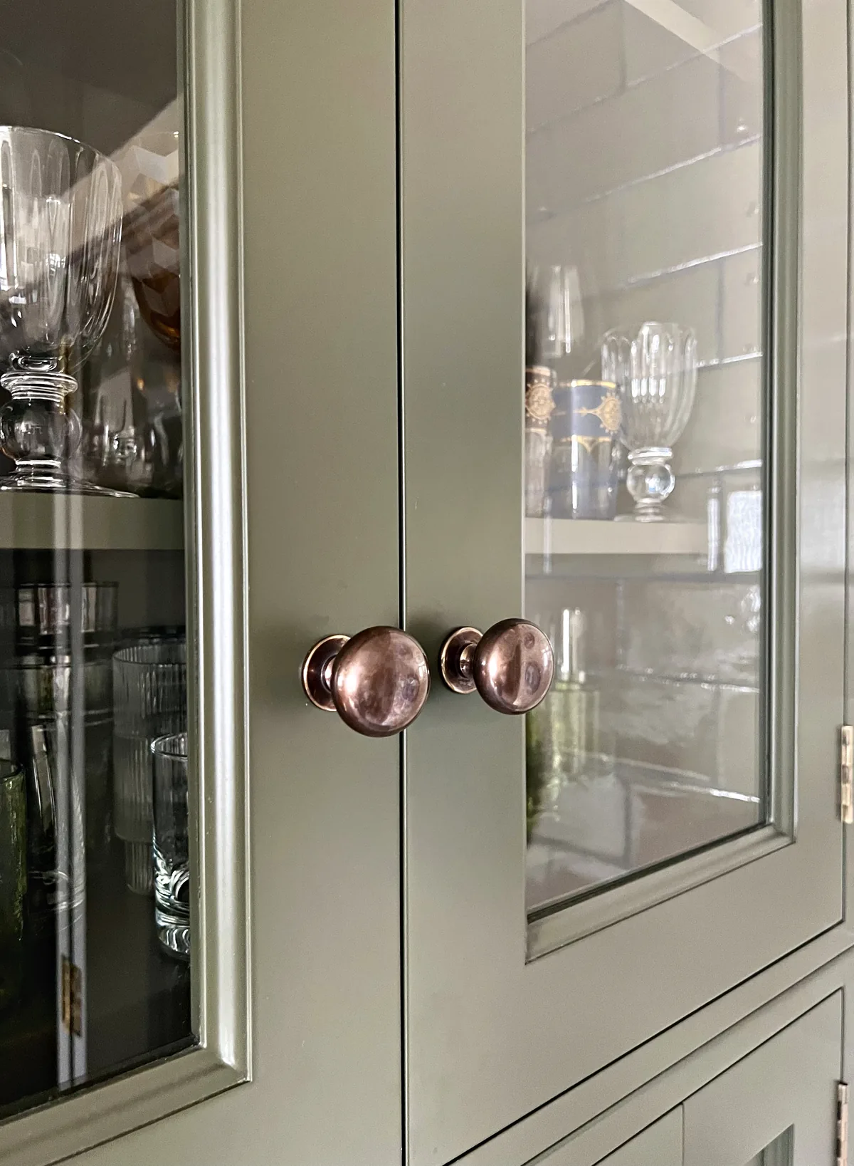 Glazed olive green dresser doors with copper knobs and glassware on the shelves behind