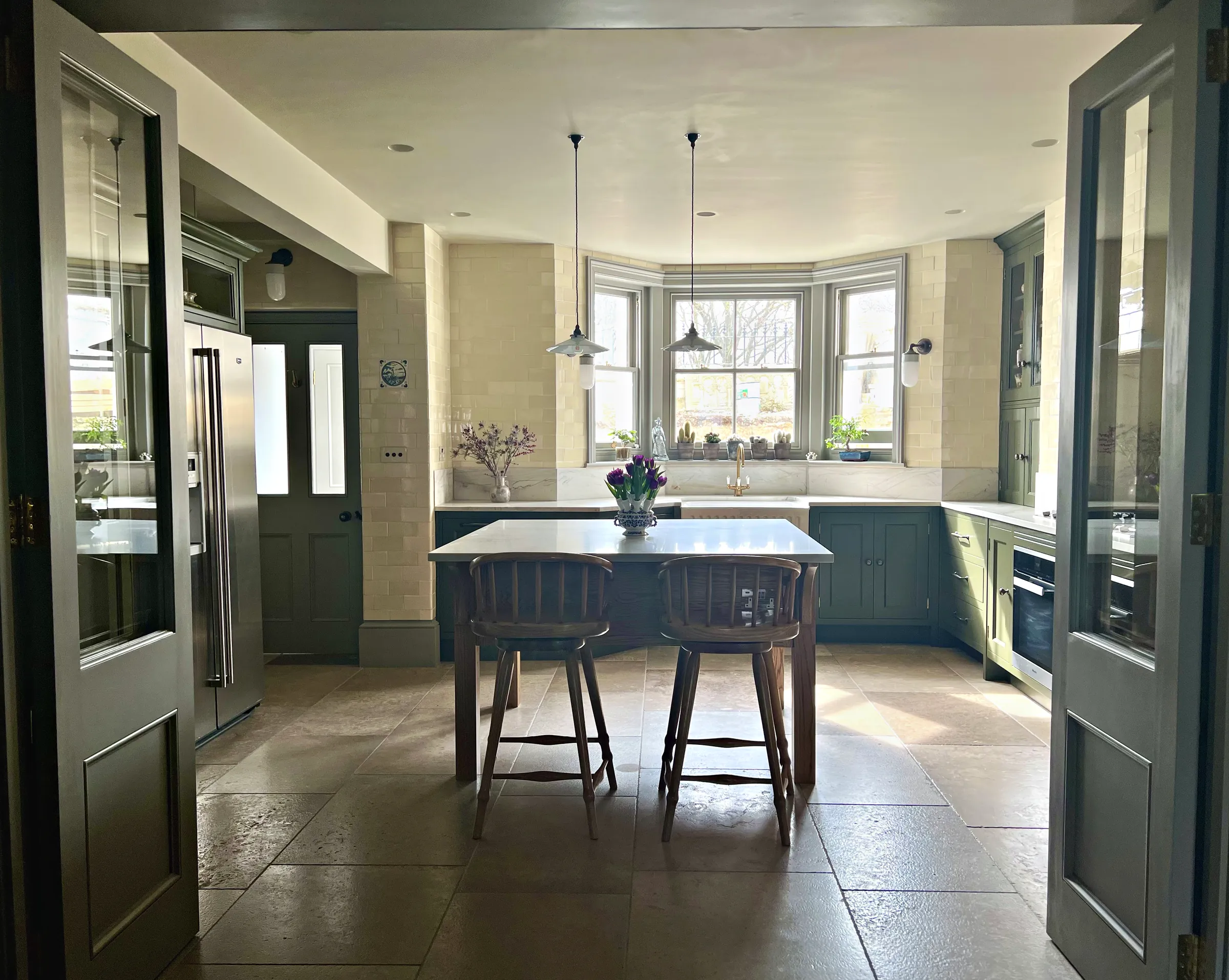 View through glazed doors into an olive green kitchen with an oak table and a bay window