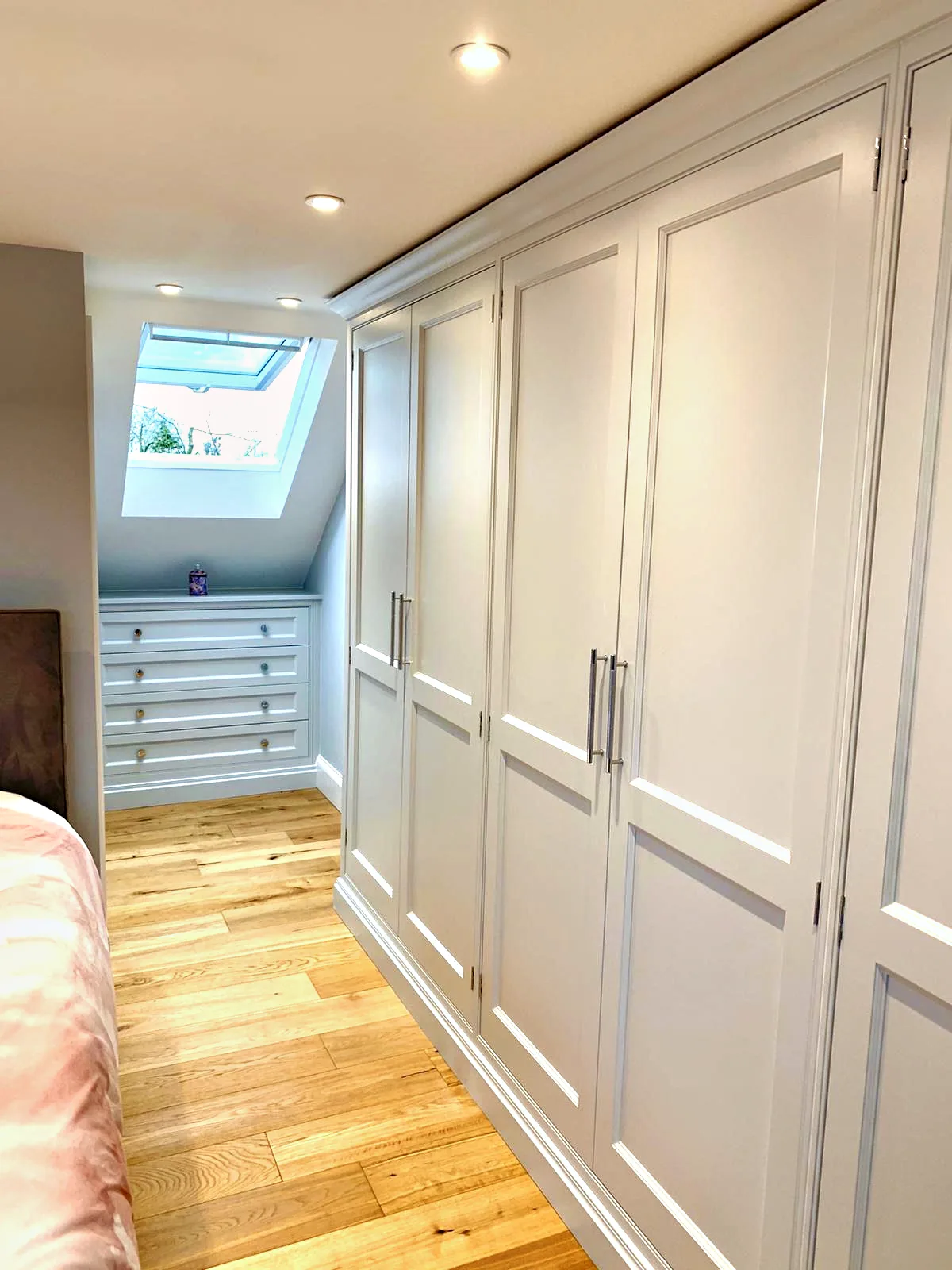 Pale fitted wardrobes under a sloping ceiling with built in drawers beneath a roof window
